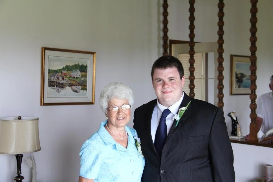 A young man in a black suit and blue tie standing next to an older woman with white hair, glasses, and a light blue dress inside a room with framed paintings on the wall.