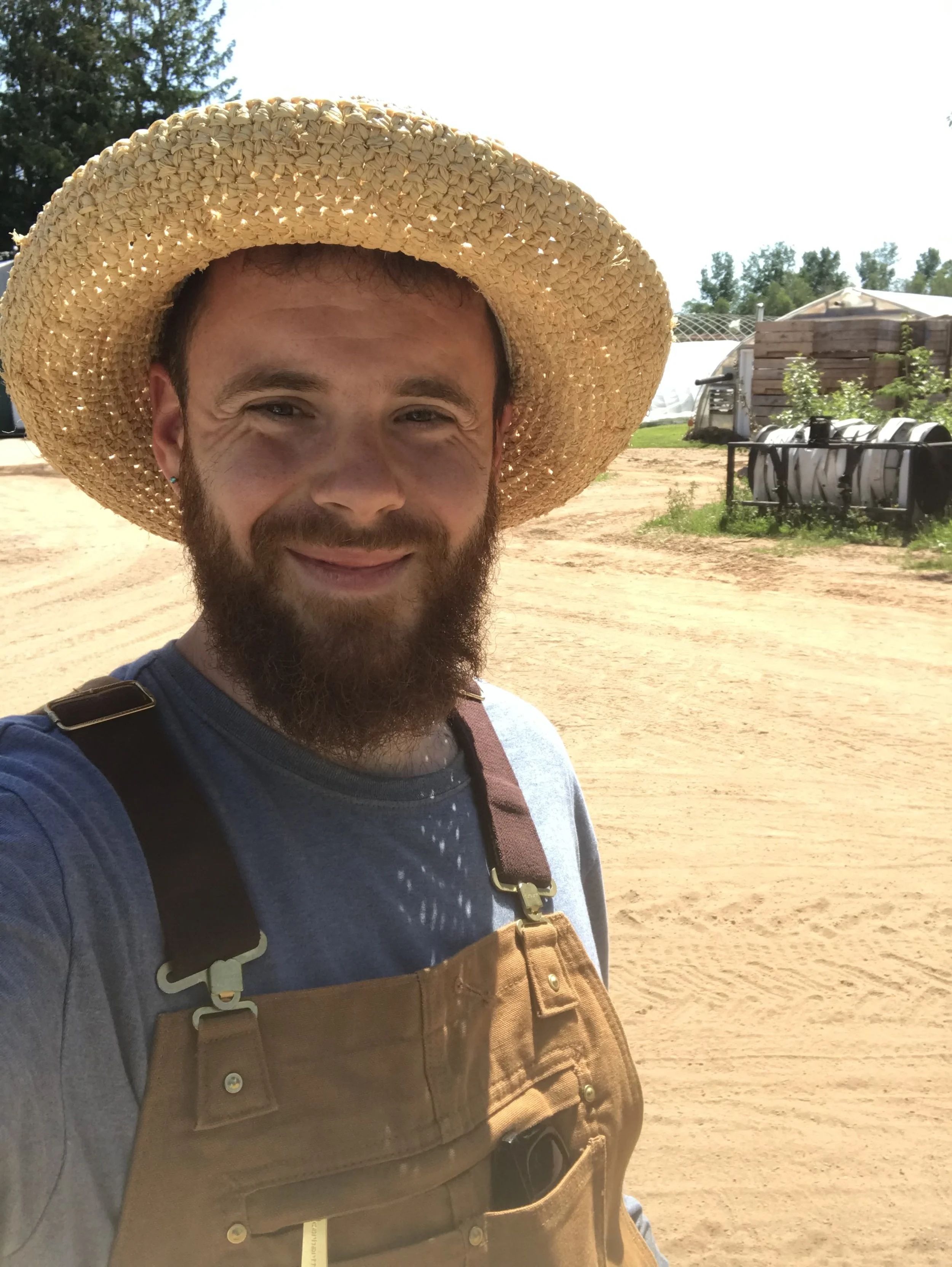 A man with a beard wearing a wide-brimmed straw hat, a gray shirt, and overalls, taking a selfie outdoors on a sunny day with a dirt path and farm equipment in the background.