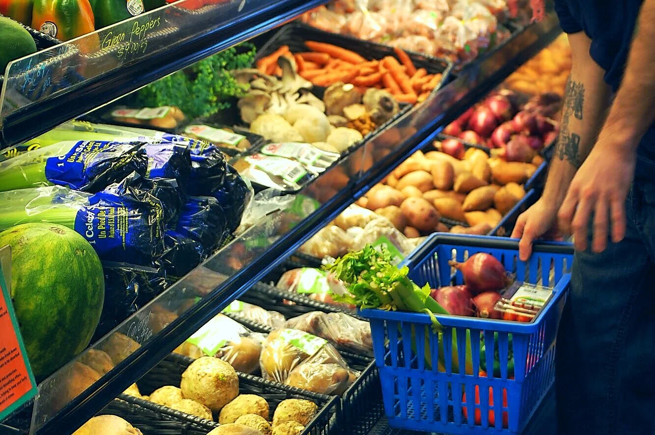 A person shopping for vegetables at a grocery store, holding a blue basket filled with red onions. The grocery shelf displays various vegetables including potatoes, carrots, mushrooms, broccoli, and others.