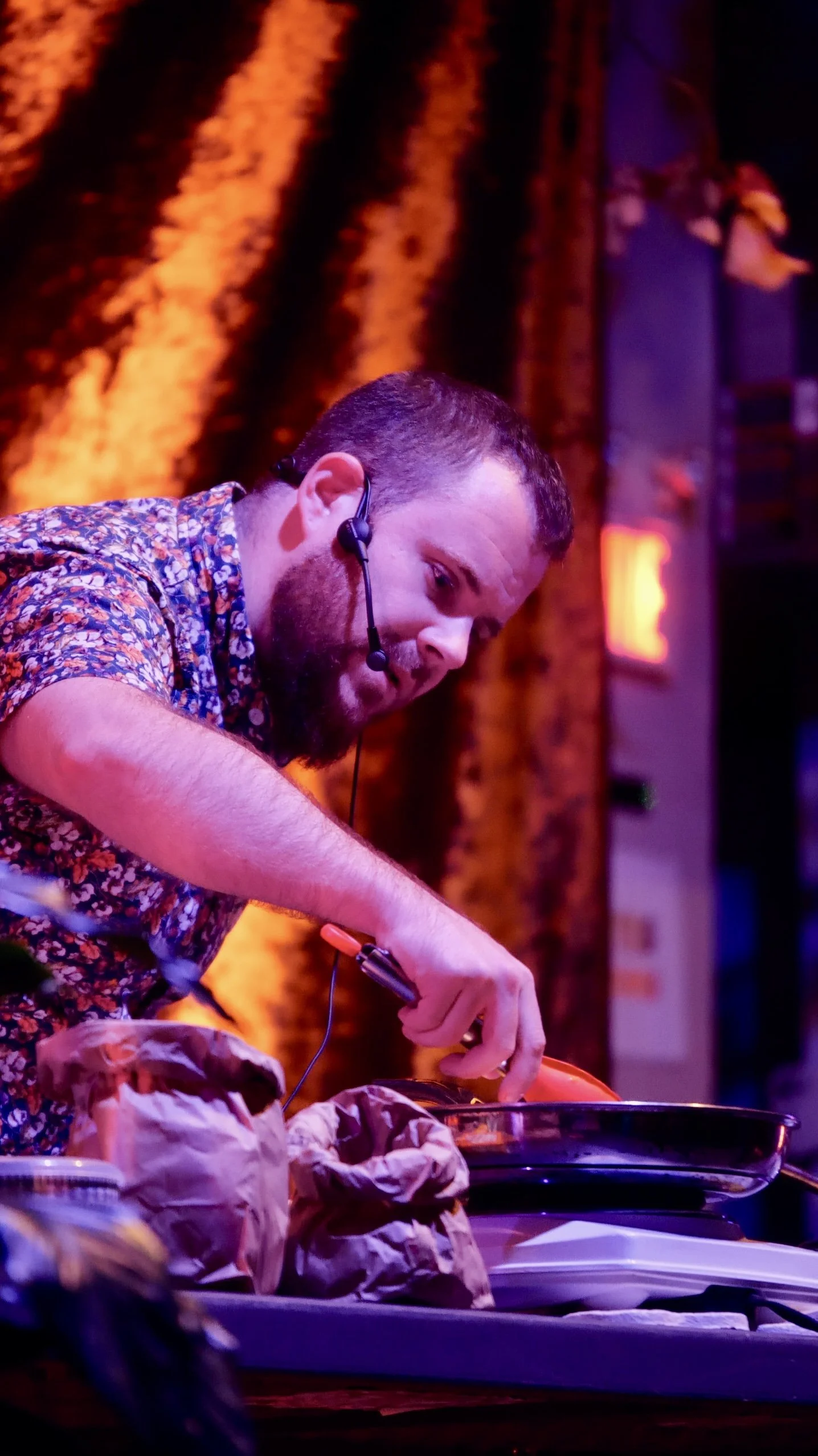 A man with a beard, wearing a headset microphone and a floral shirt, is cooking or preparing food on a stove in a dimly lit environment with warm, colorful lighting.