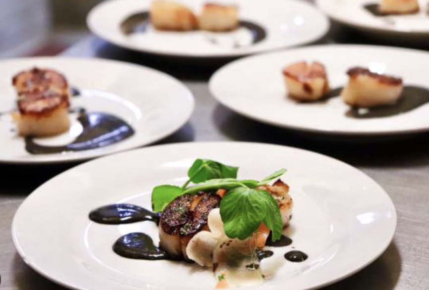 A plated gourmet dish with a seared scallop, fresh green herbs, and drizzled sauce, surrounded by several other similar plates of scallops in the background.