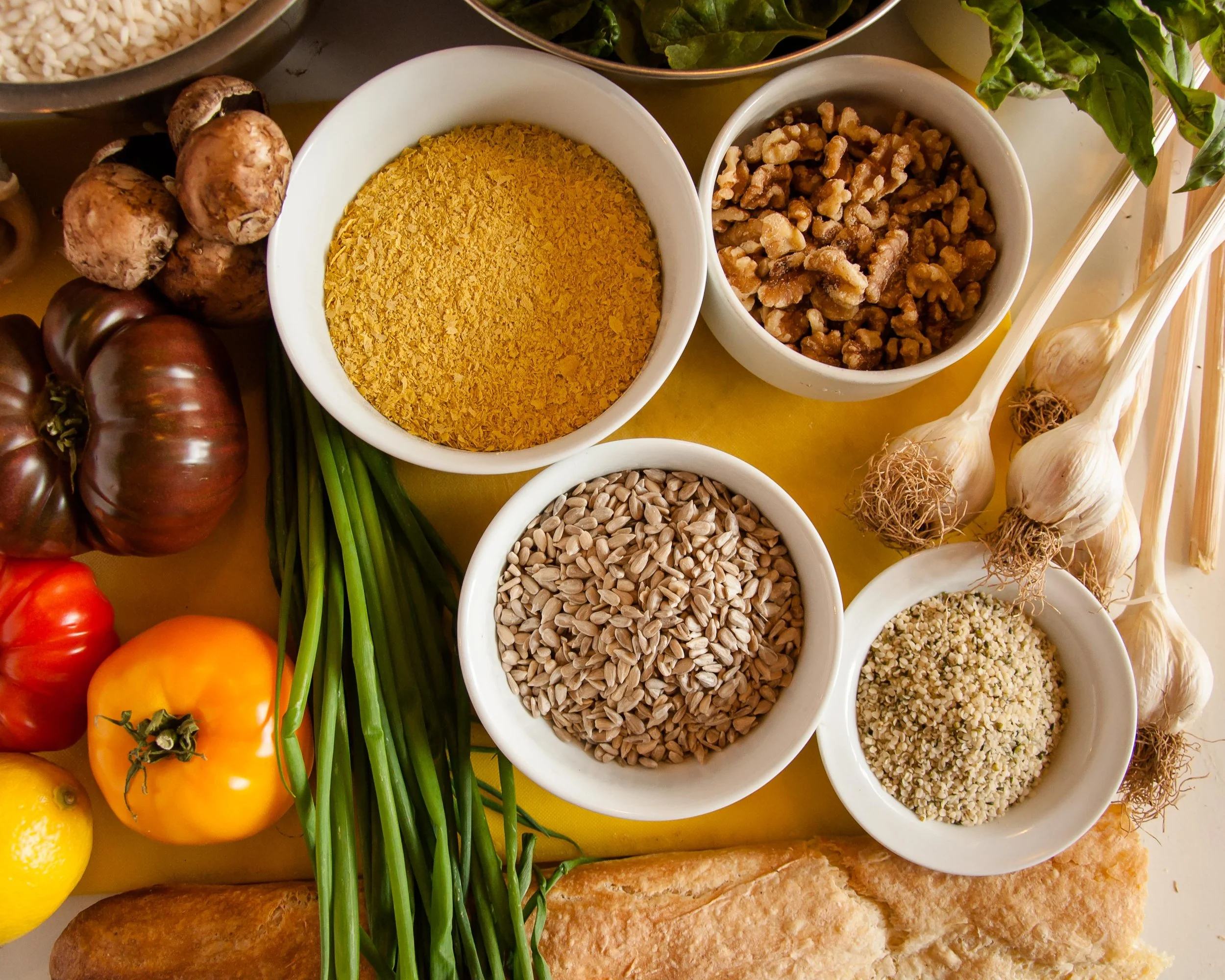 Fresh vegetables and spices, including garlic, tomatoes, lemon, chives, and various bowls of seeds and seasonings on a yellow surface.