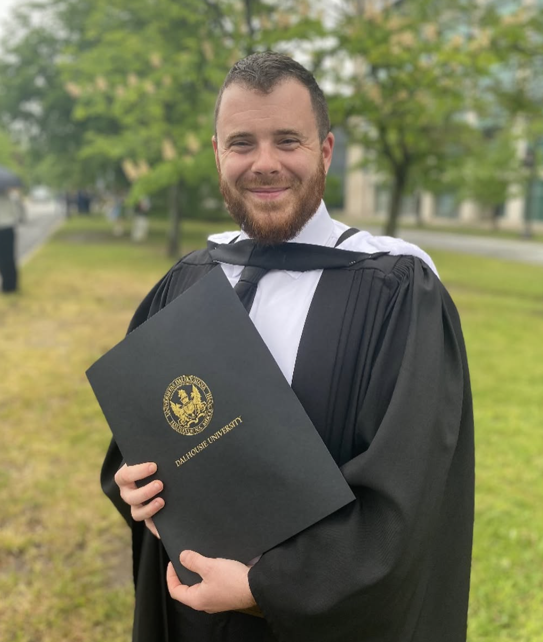 A man in a graduation gown holding a diploma from Dalhousie University, smiling outside with trees and a building in the background.