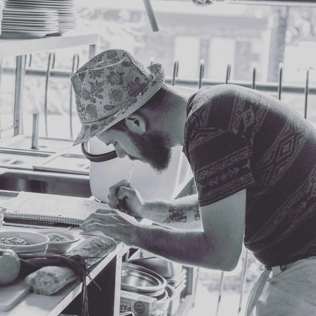 A man with tattoos, a beard, wearing a patterned shirt, and a bucket hat, preparing food in a kitchen, using a spray bottle.