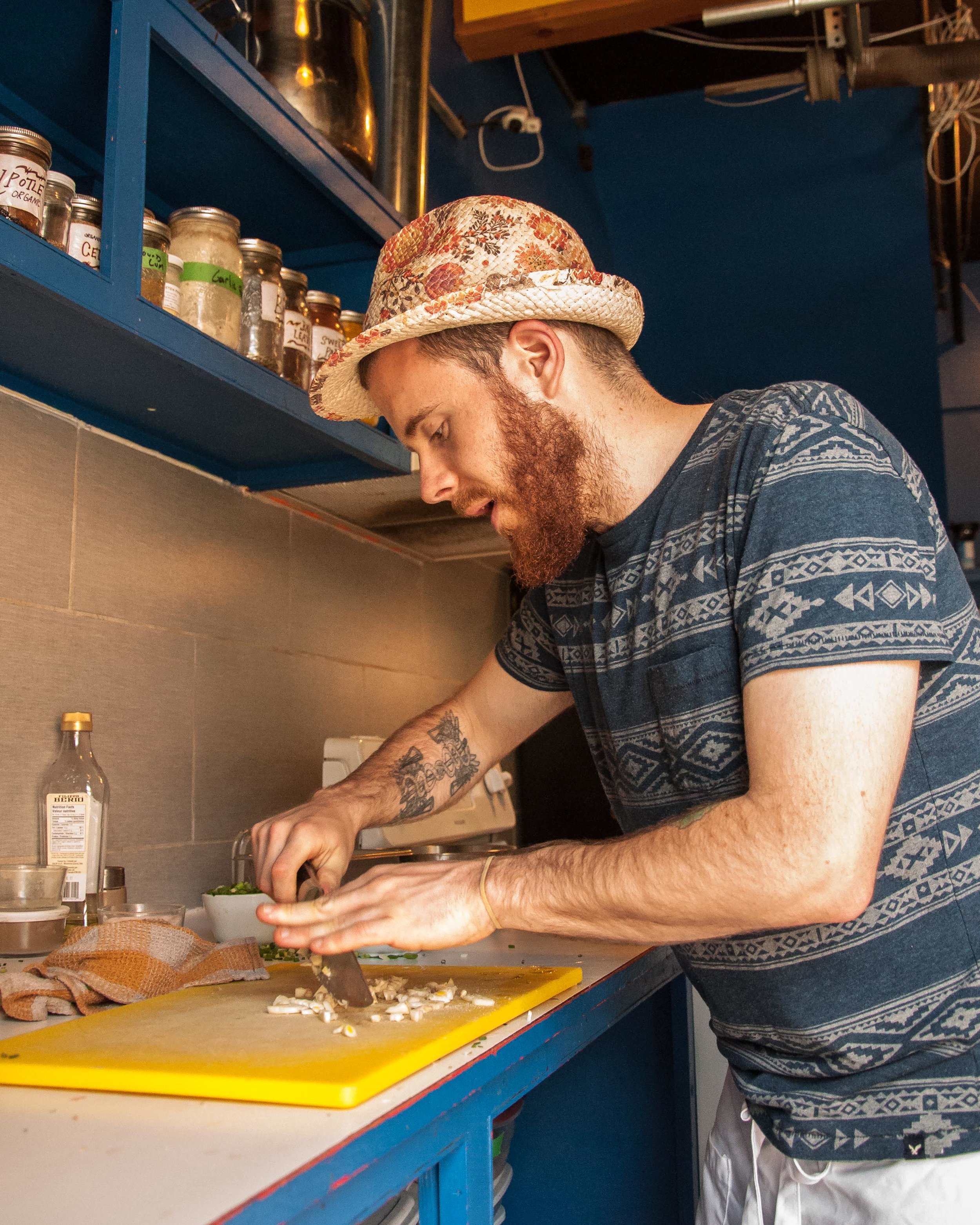 Man wearing a floral hat and patterned T-shirt chopping vegetables in a kitchen with jars on the blue shelf behind him.