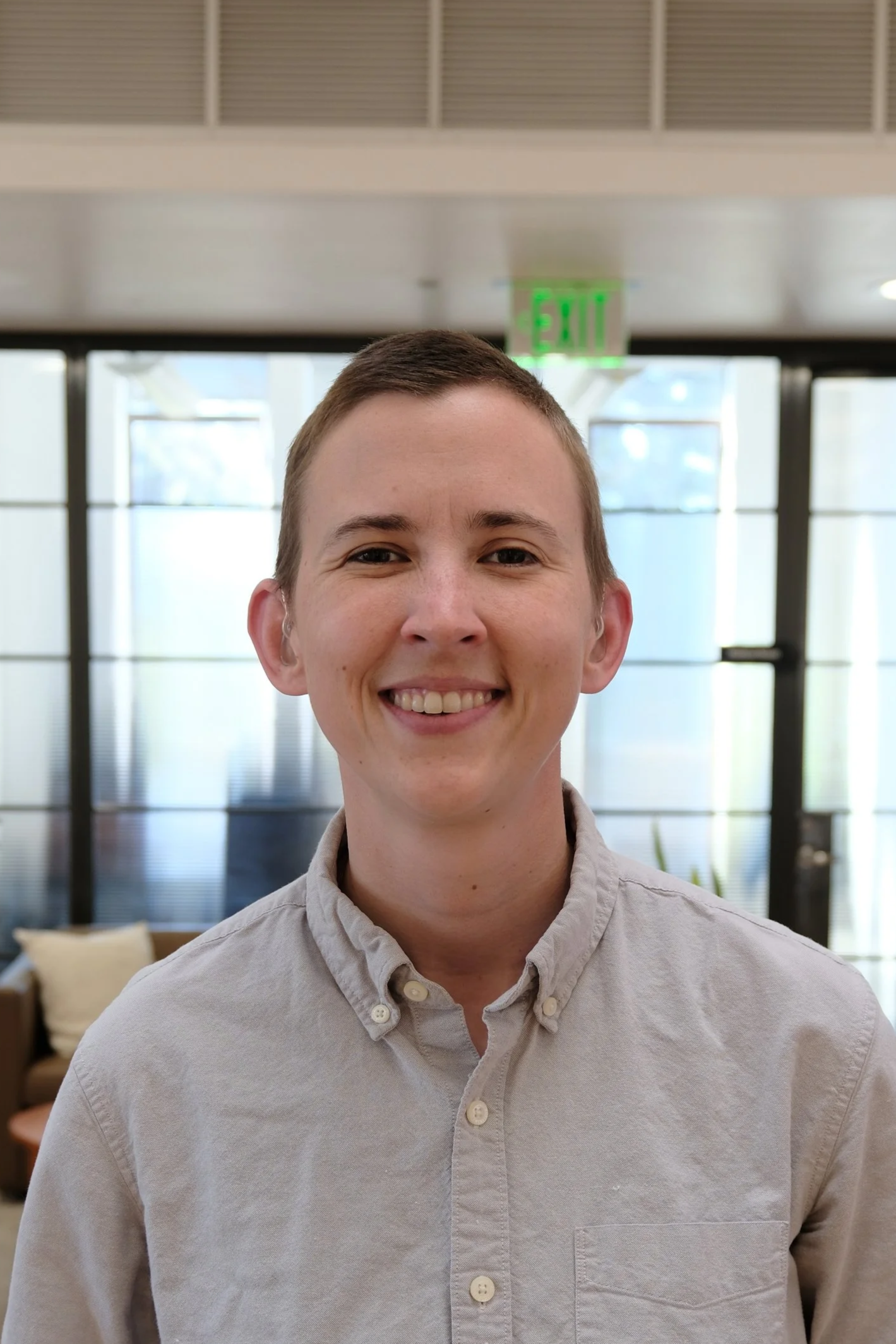 A smiling young man with short hair wearing a light-colored button-up shirt, standing indoors with large windows and an "EXIT" sign in the background.