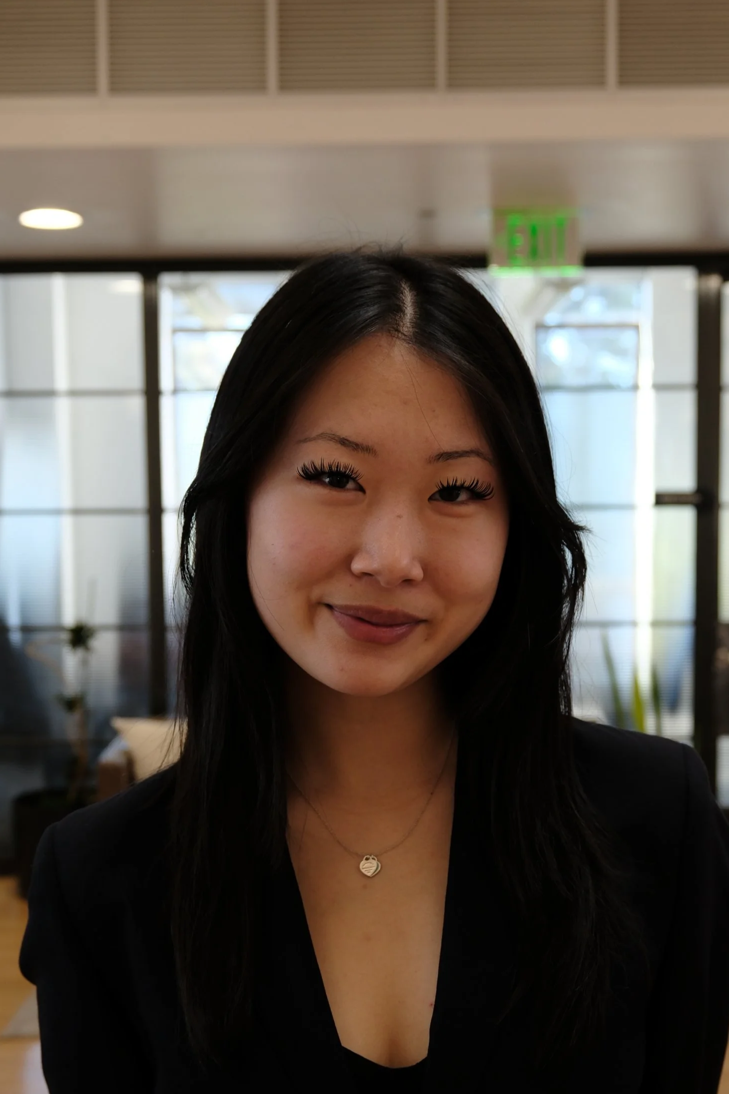 Portrait of a young woman with long black hair smiling, wearing a black blazer and a necklace, inside a modern building with large windows and an exit sign in the background.