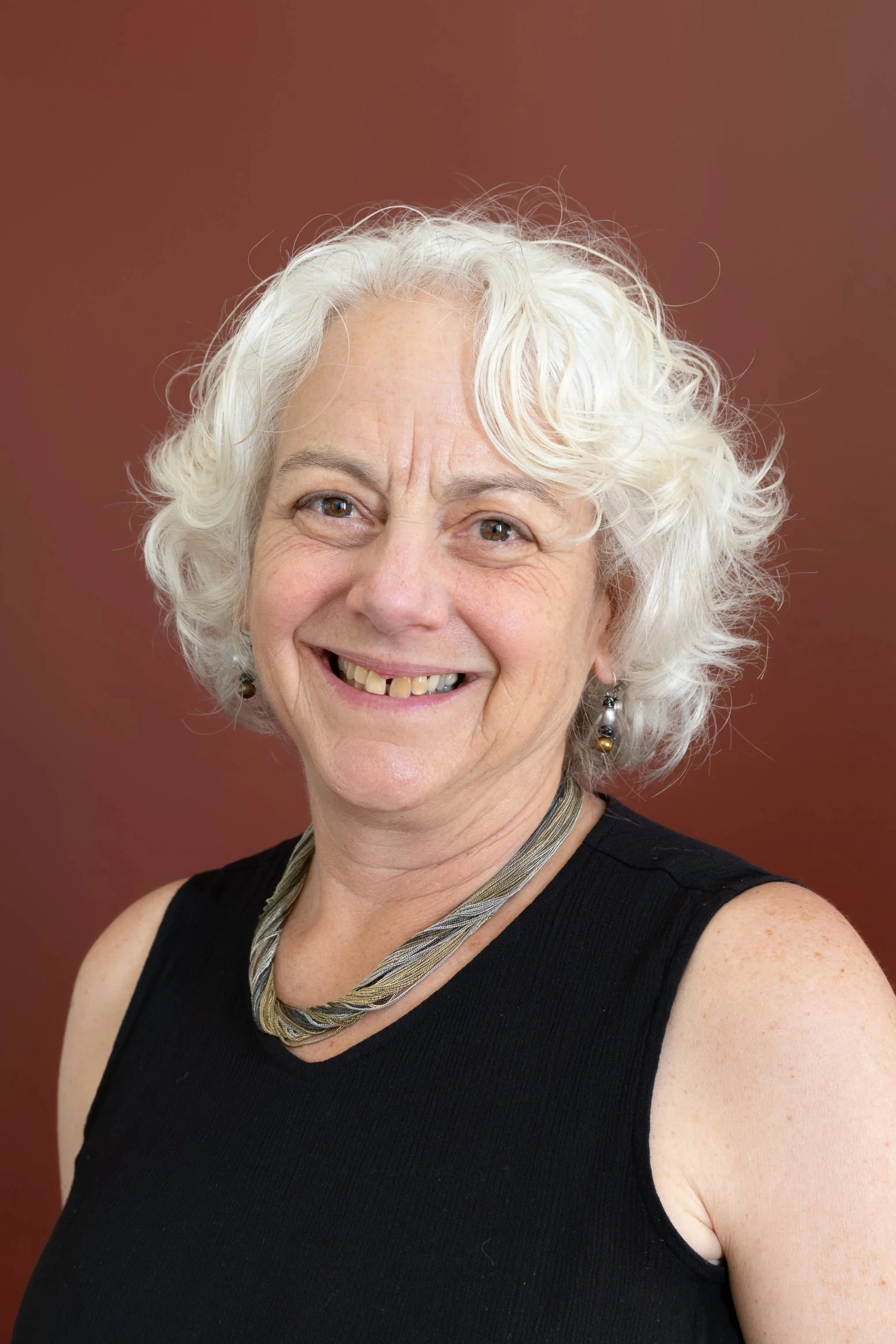Portrait of an elderly woman with curly white hair, smiling, wearing a black sleeveless top, a twisted necklace, and earrings, against a solid brown background.