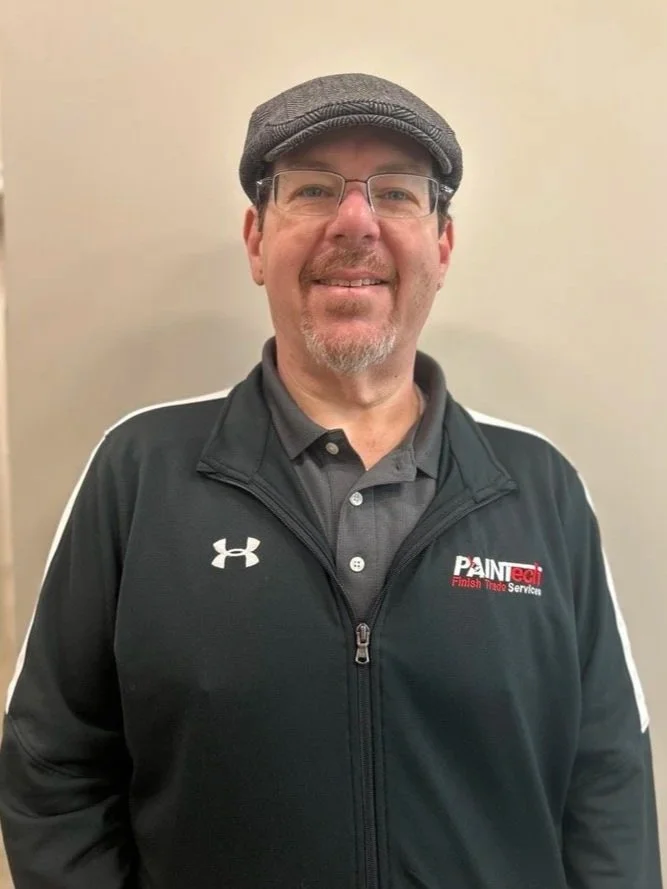 A smiling man wearing glasses and a gray flat cap, dressed in a black Under Armour jacket with a 'Painttech Finish Trade Services' logo, standing against a beige wall.