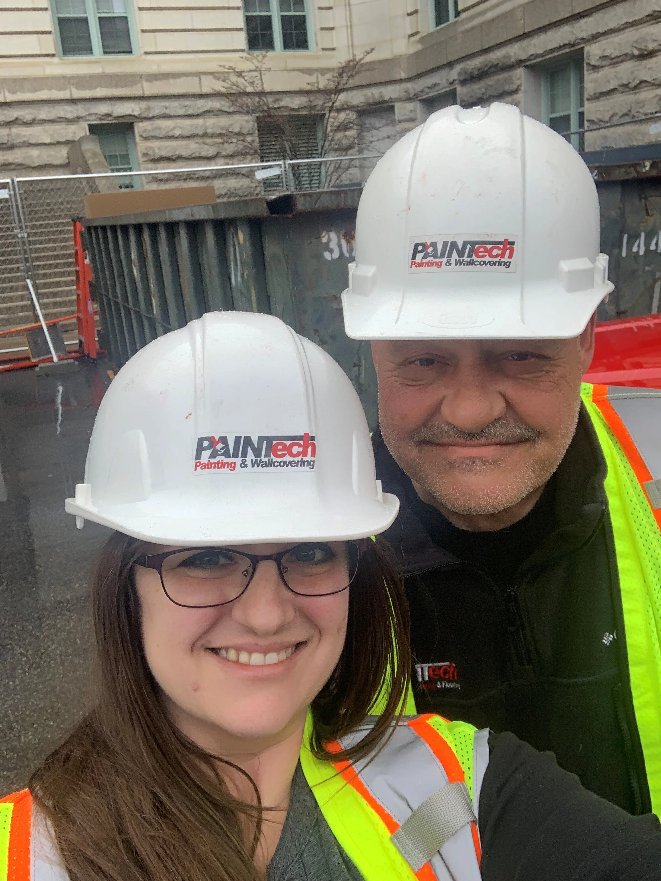 Two construction workers, a man and a woman, wearing safety helmets with a company logo and high-visibility safety vests, taking a selfie at a construction site with a building and a dumpster in the background.