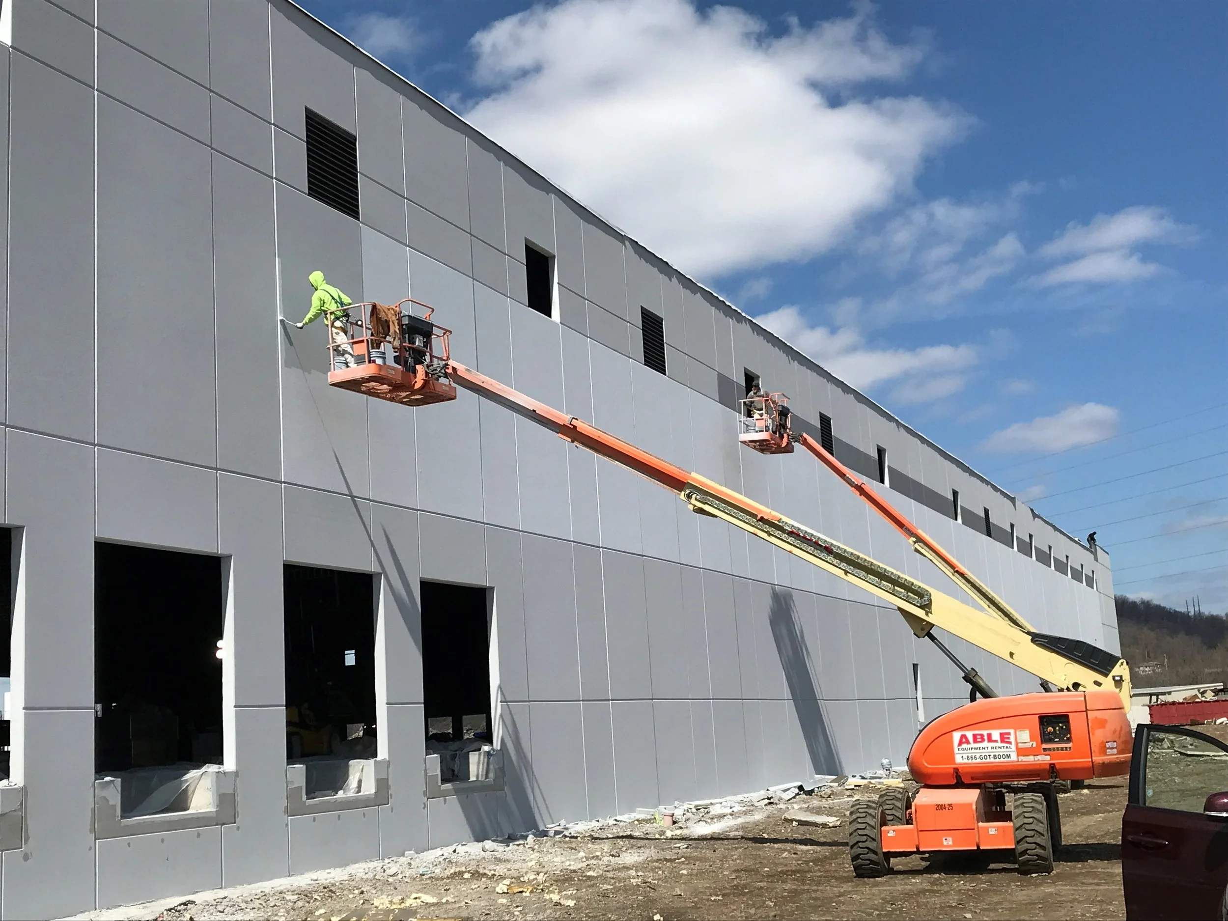 Construction workers are on an elevated work platform installing exterior wall panels on a modern building. The building has large windows and is gray with rectangular panels. The sky is partly cloudy.