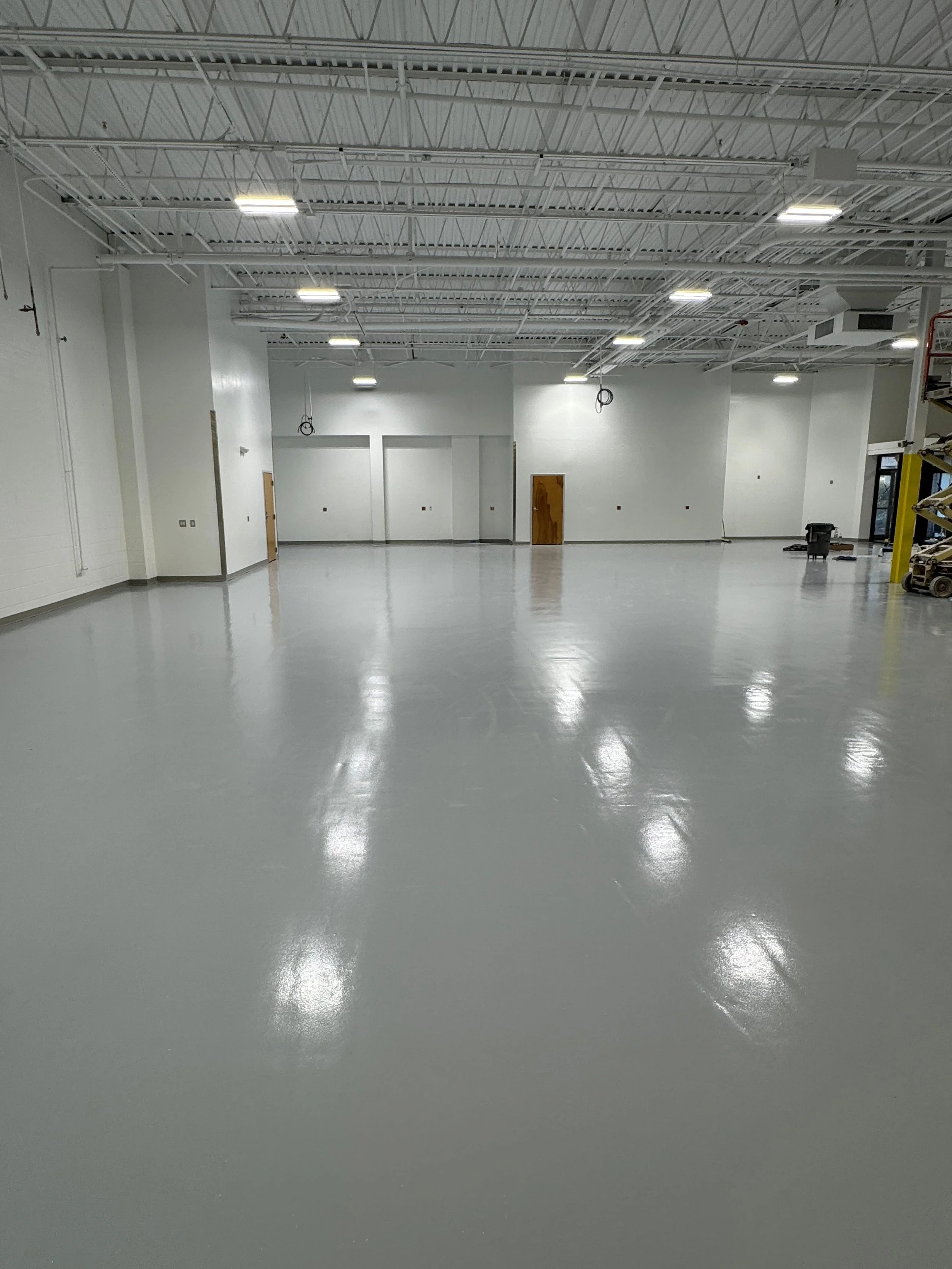 An empty indoor space with white walls, a high ceiling with exposed metal beams, bright recessed lighting, and a smooth, polished gray floor. There are some construction or maintenance tools and equipment along the right wall.