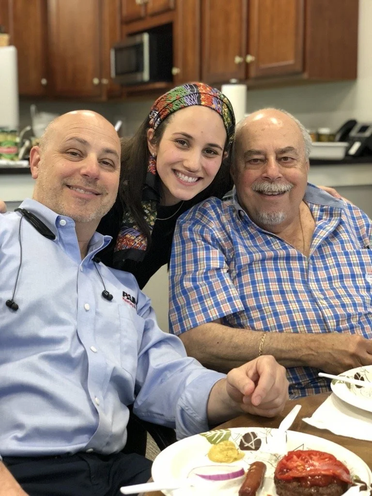 Three smiling people sitting at a table with plates of food, in a kitchen setting.