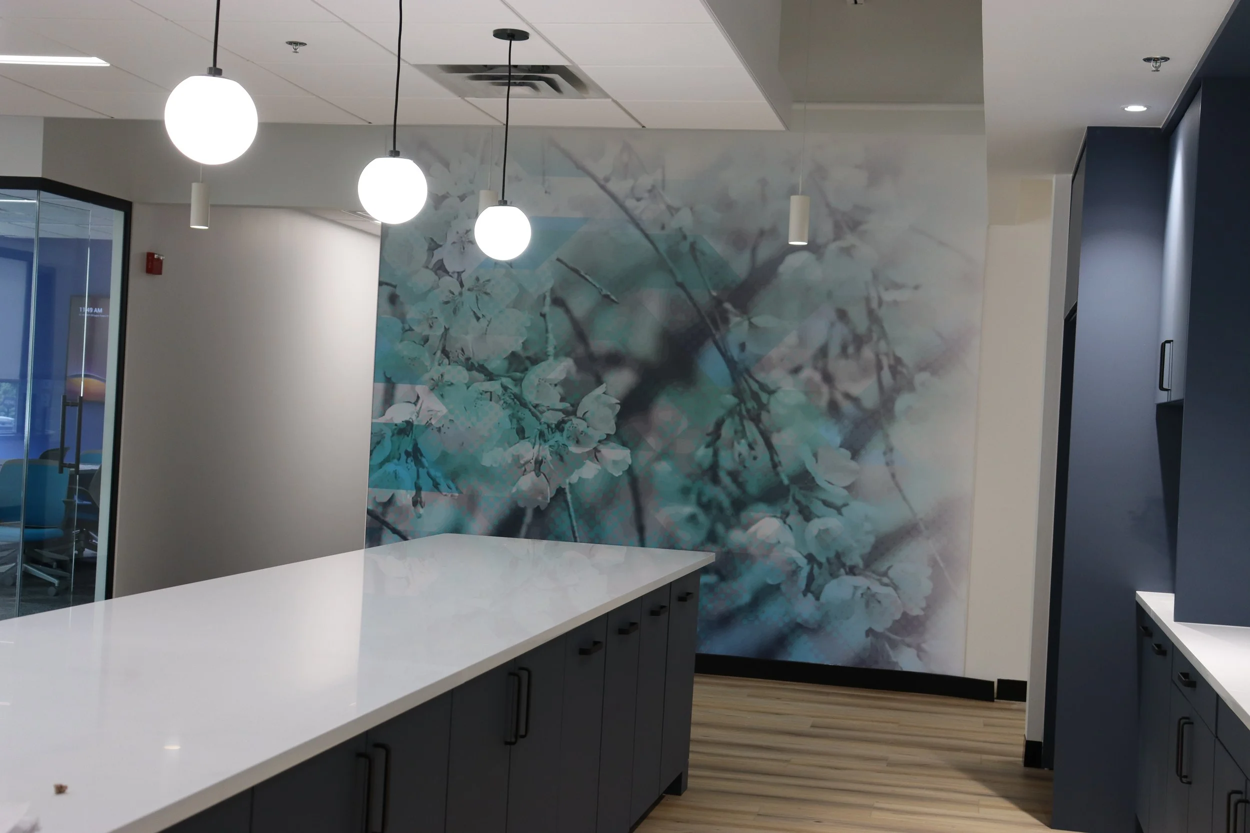 Interior view of a modern office with a white countertop, black cabinets, hanging spherical lights, a mural of cherry blossoms on the wall, and a glass door leading to a meeting room.