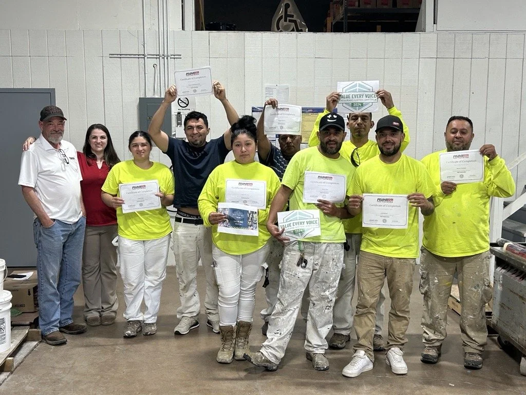 Group of ten individuals, some wearing yellow shirts, holding certificates, standing in a warehouse or factory setting.