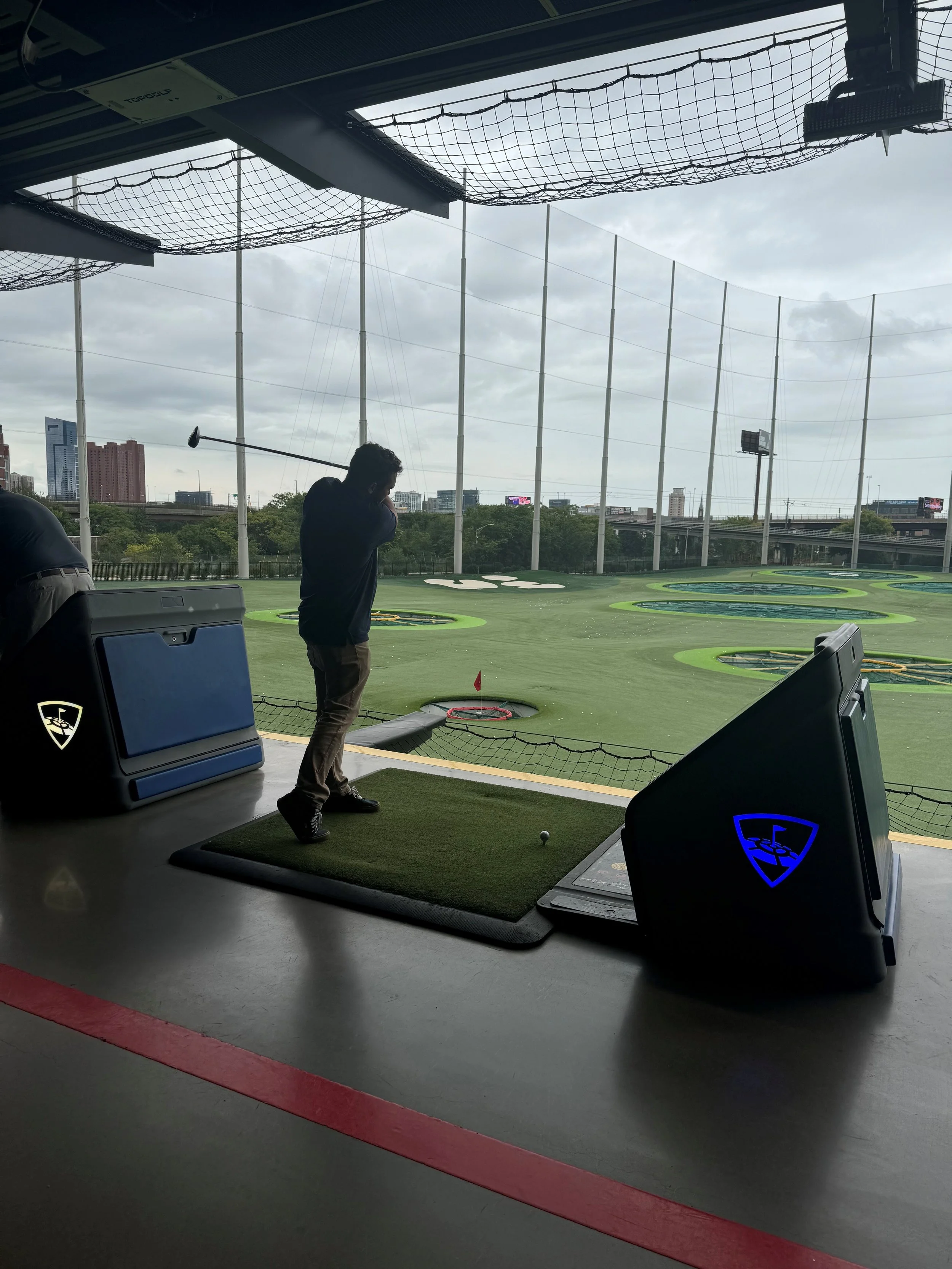 A person practicing golf at an indoor driving range, preparing to hit the golf ball with a golf club. The range has multiple targets on the green field outside, with a cityscape and cloudy sky in the background.