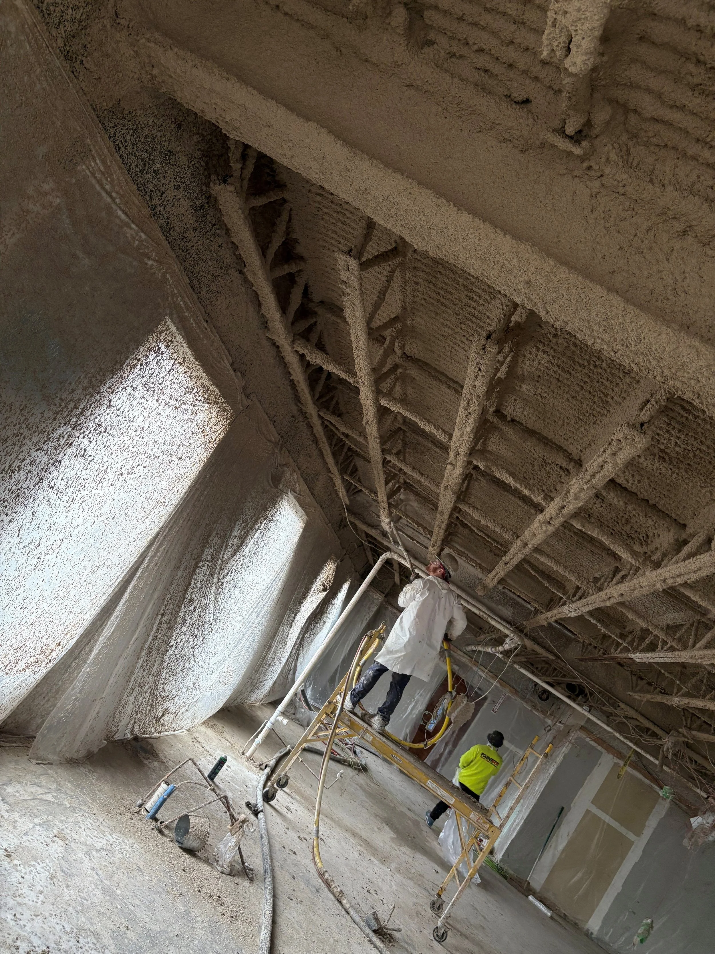 Construction workers applying spray foam insulation to the ceiling of a building under construction.