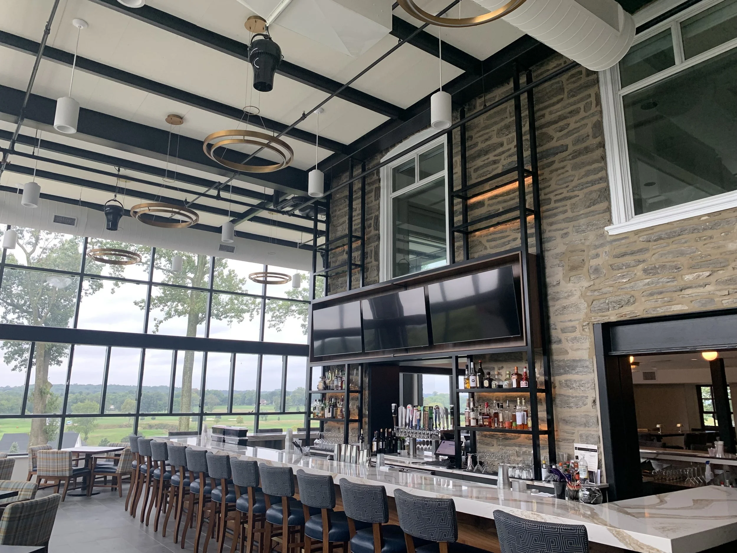 Modern indoor bar with large windows, stone wall, and circular ceiling lights, overlooking green landscape.