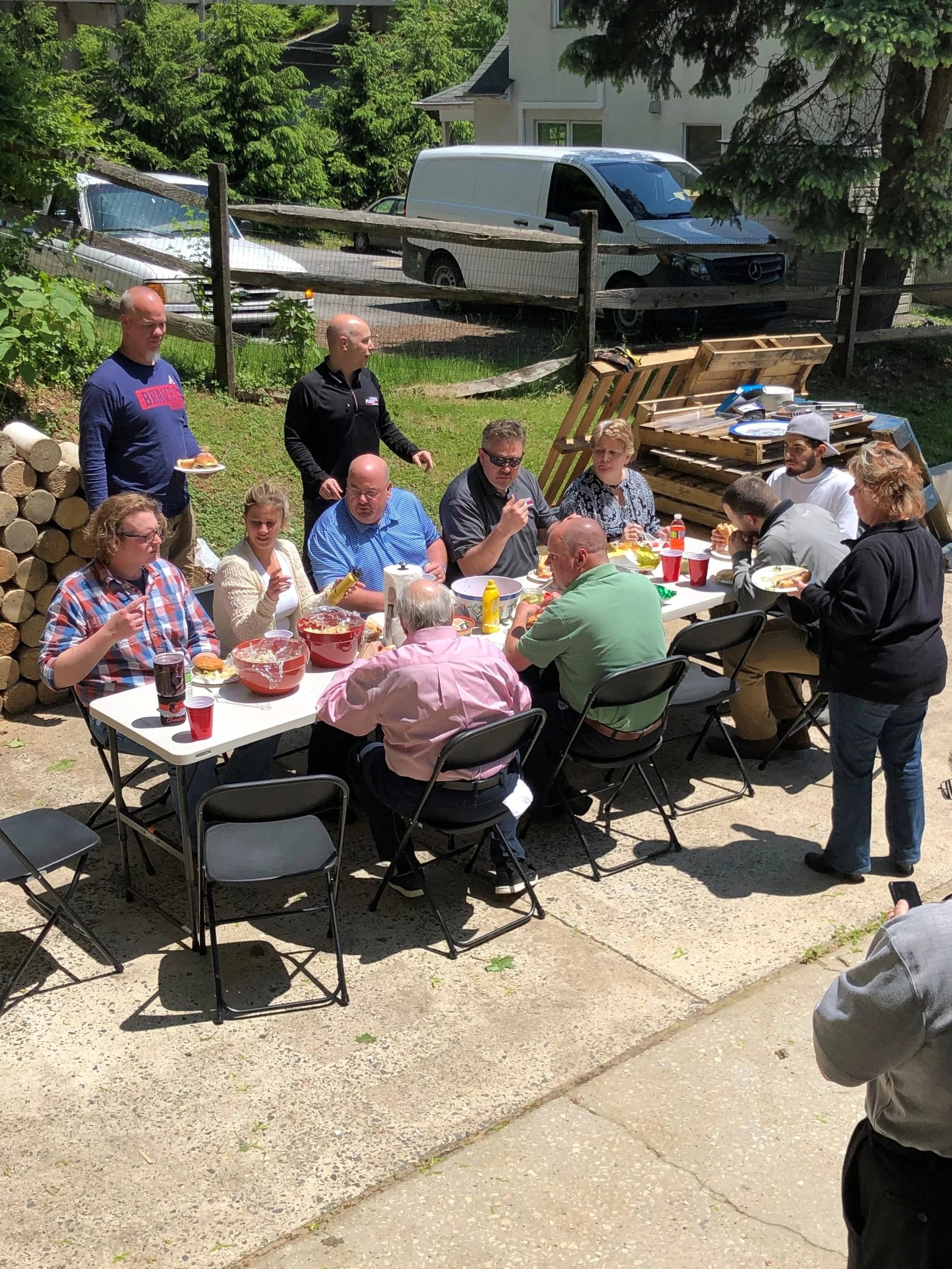A group of people gathered outdoor around a table, eating and socializing, with wood logs and parked vehicles in the background.