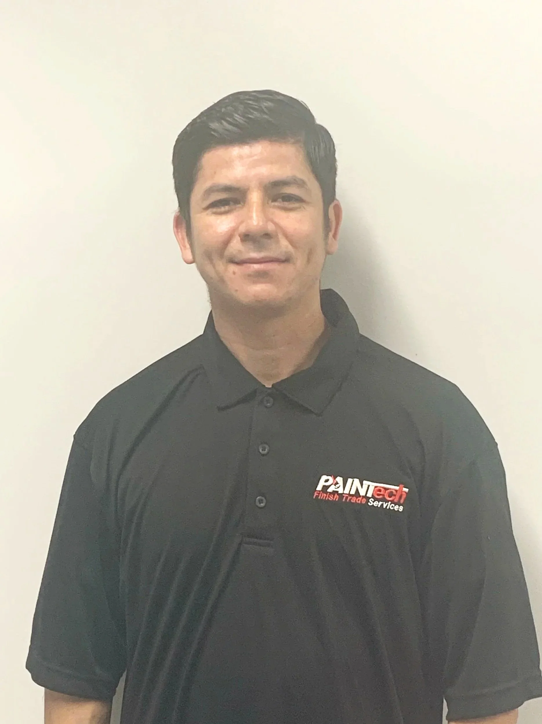 A man standing against a plain white wall, wearing a black collared shirt with a logo that reads "PAINtech Finish Trade Services."
