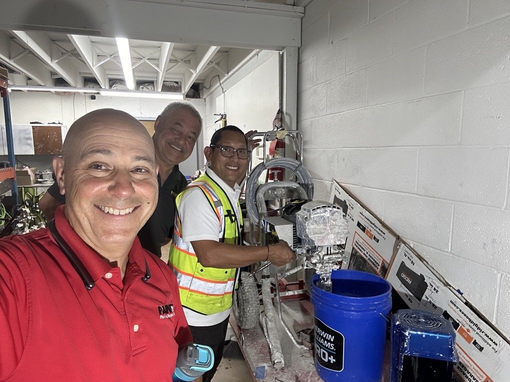 Four men, three in safety vests and one in a red shirt, smiling and taking a selfie in a workshop with machinery and tools on workbenches.