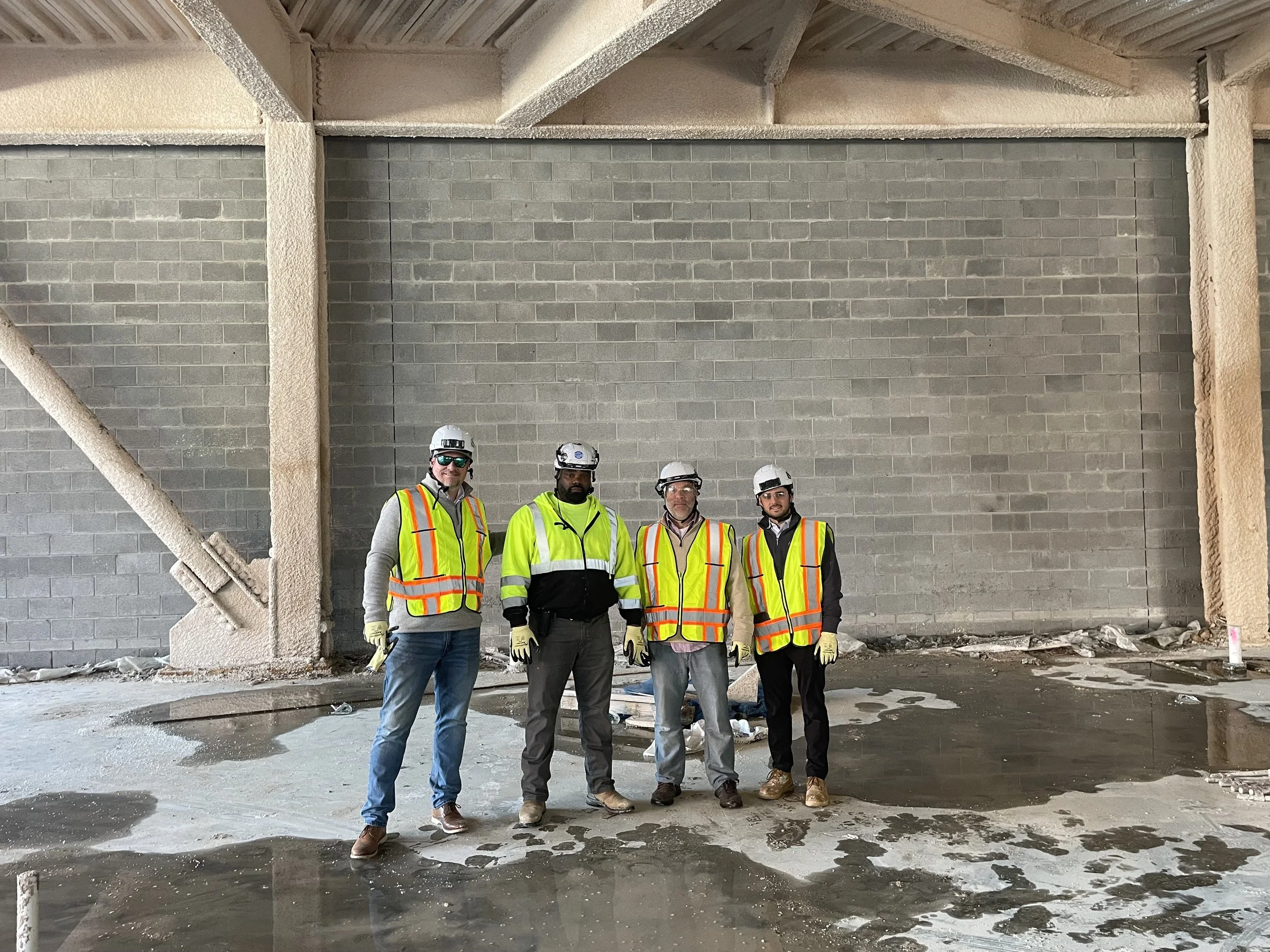 Four construction workers standing in front of a brick wall inside a building under construction, wearing safety vests and helmets.