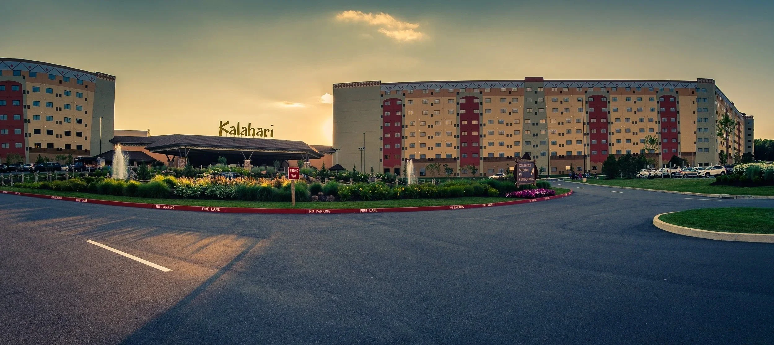Exterior of a large hotel named Kalahari, with a landscaped driveway, flowers, fountains, and parking lot, during sunset.
