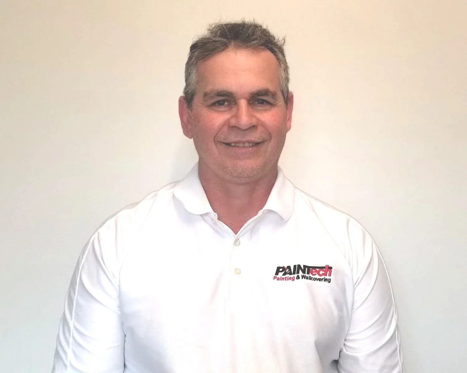 A middle-aged man with gray hair, wearing a white punctual shirt with a logo that reads 'PAINtech Painting & Wallcovering,' standing against a plain, light-colored wall and smiling at the camera.