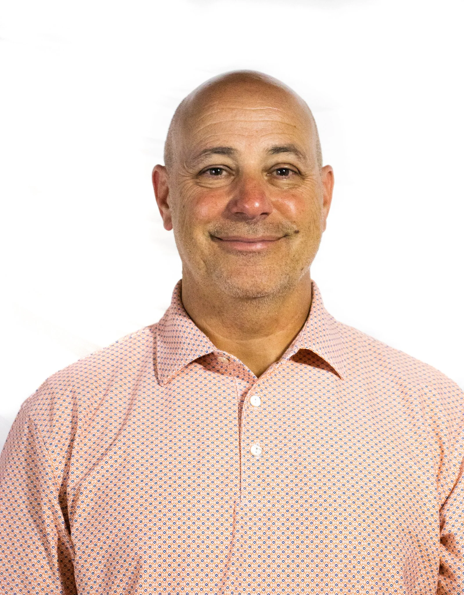 A smiling man with a bald head wearing a peach-colored collared shirt with a small dot pattern, against a white background.