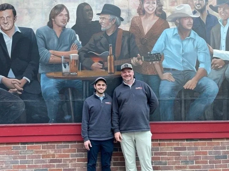 Two men standing in front of a large mural of people at a bar, including a man with a guitar and others sitting around, painted on a brick wall.