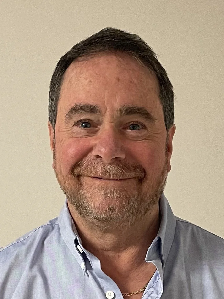 Close-up photo of a smiling middle-aged man with dark hair, a beard, wearing a light blue button-up shirt against a plain beige wall.