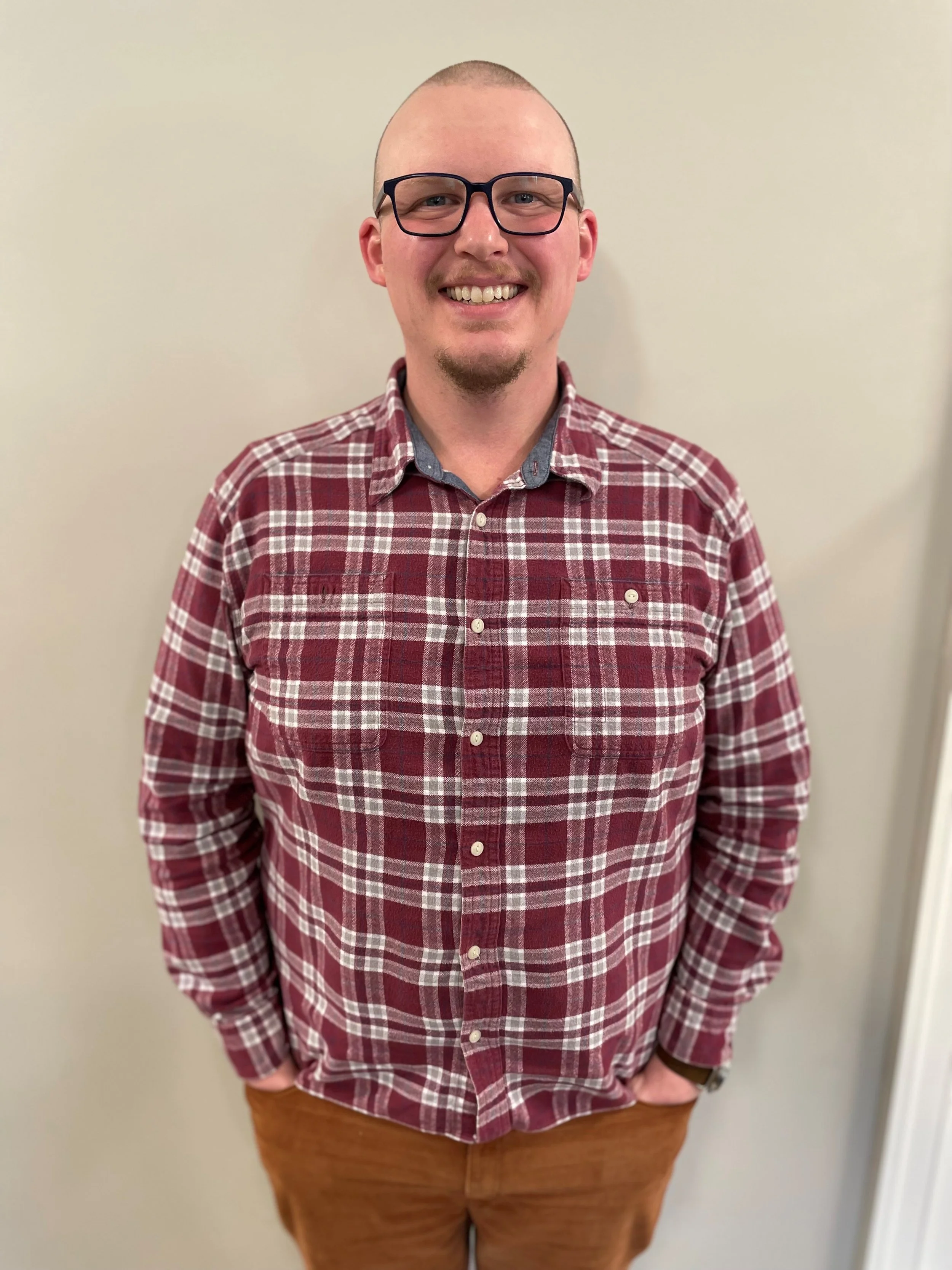 A man with glasses, a goatee, and a buzz cut head, dressed in a red plaid shirt and brown pants, smiling and standing against a plain beige wall.