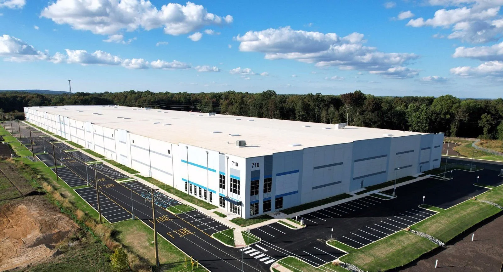 Large modern industrial warehouse building with multiple parking lots, some designated for handicap parking, under a bright blue sky with scattered clouds.