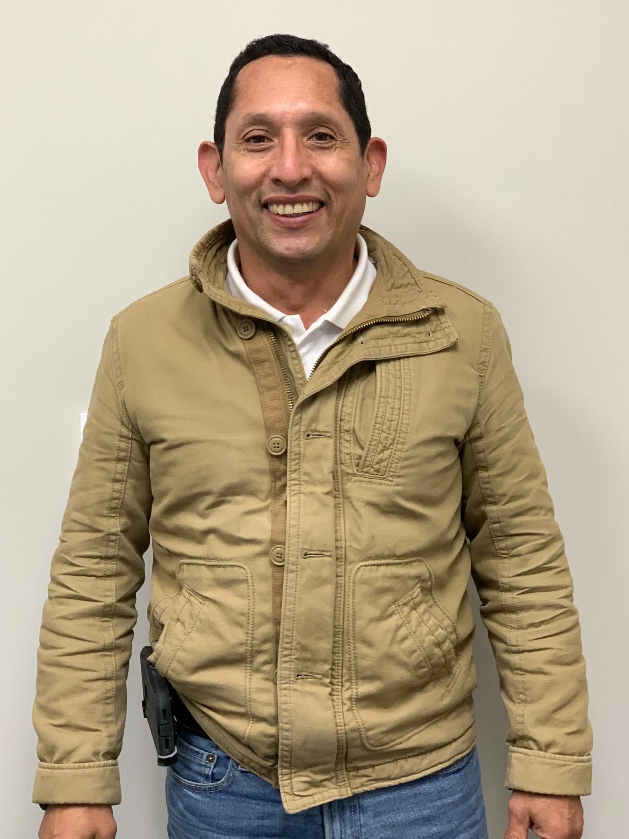 A man wearing a tan jacket over a white shirt, standing against a plain white wall, smiling.