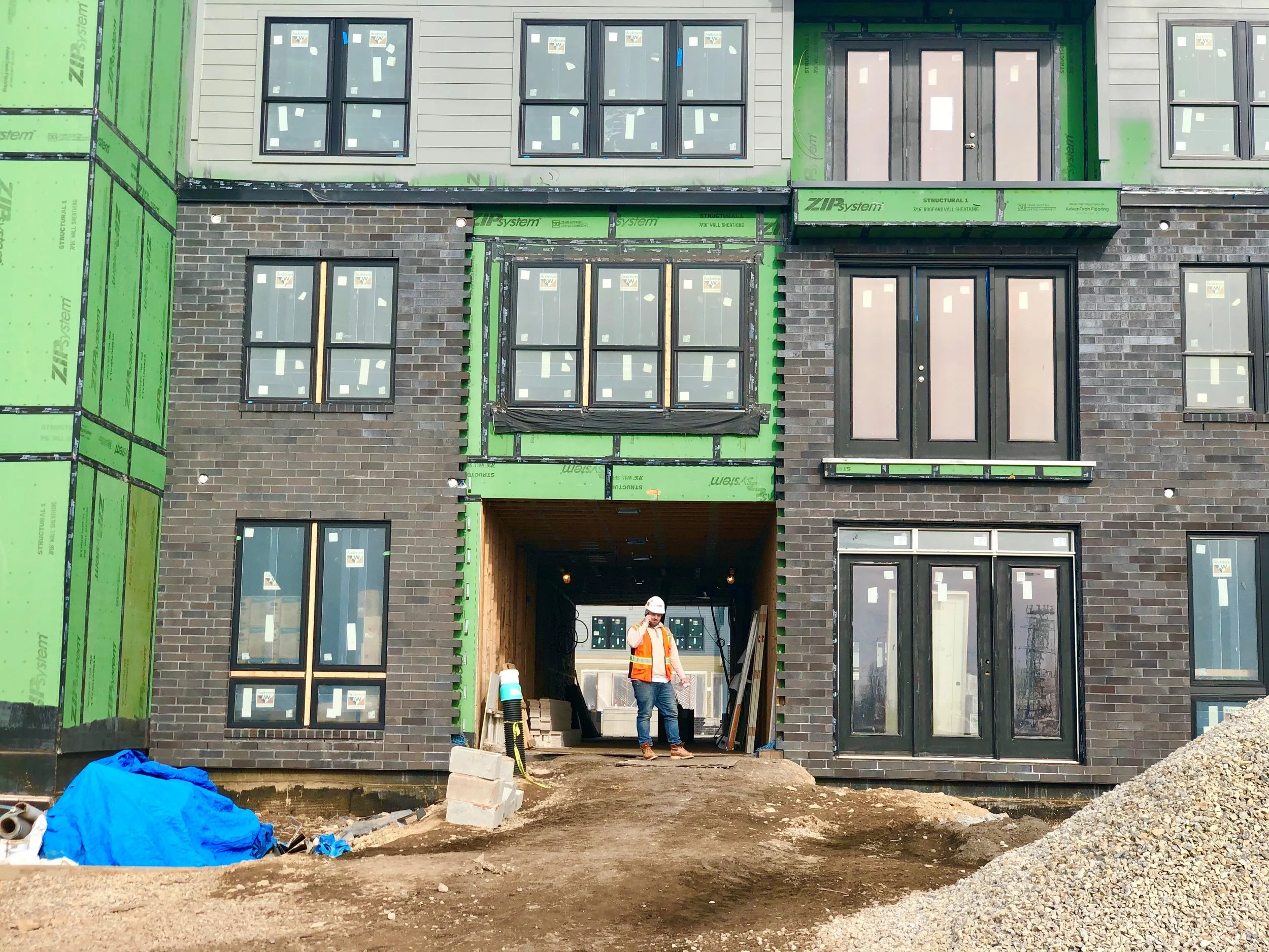 Construction workers at a building under construction, with a partially finished brick facade, green sheathing, and multiple windows installed, as well as construction materials and equipment on site.