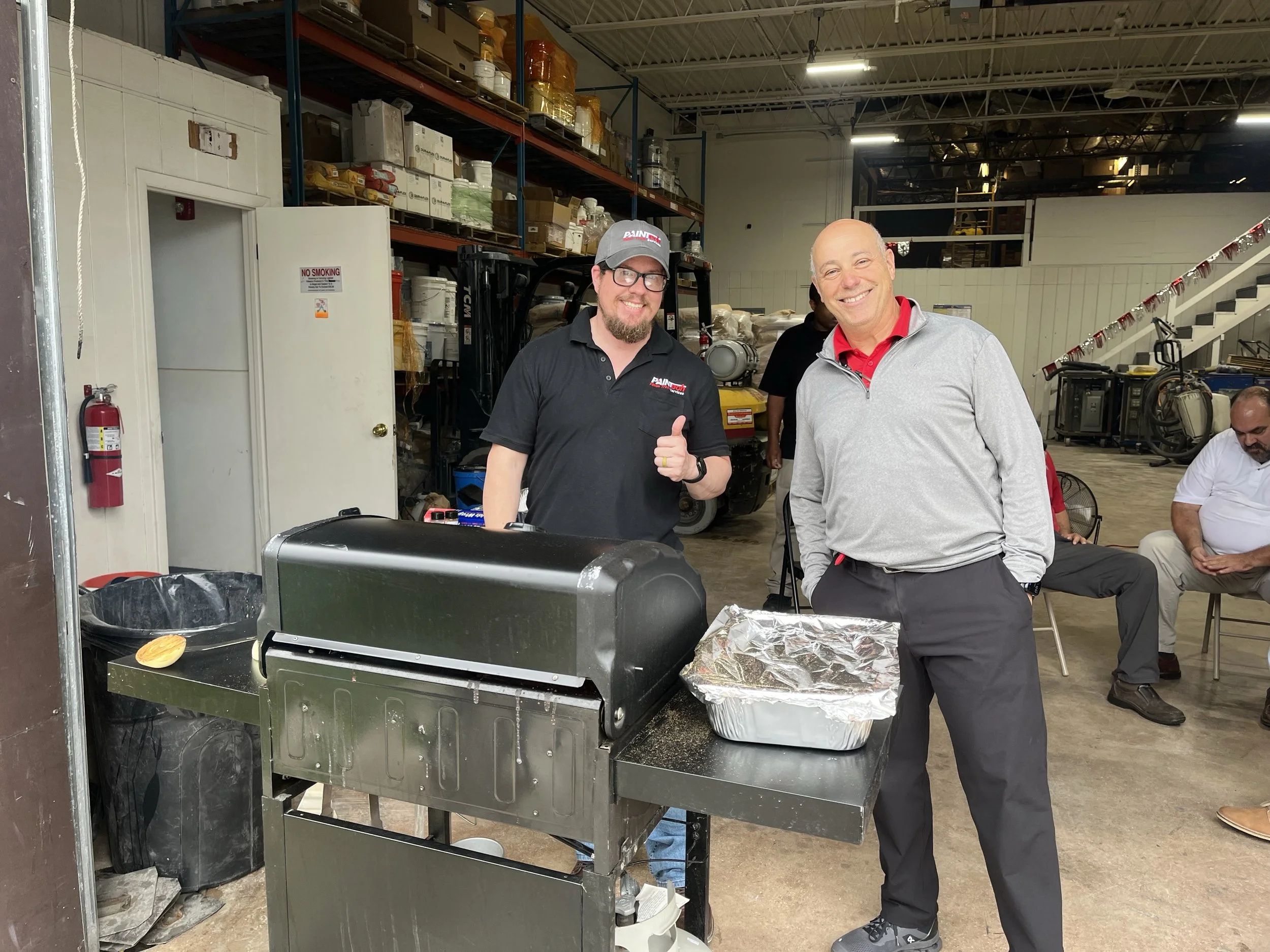Two men smiling at a gathering in a warehouse or industrial space, one giving a thumbs-up. There is a grill with food and foil in the foreground, shelves with boxes and equipment in the background, and other people seated or standing around.