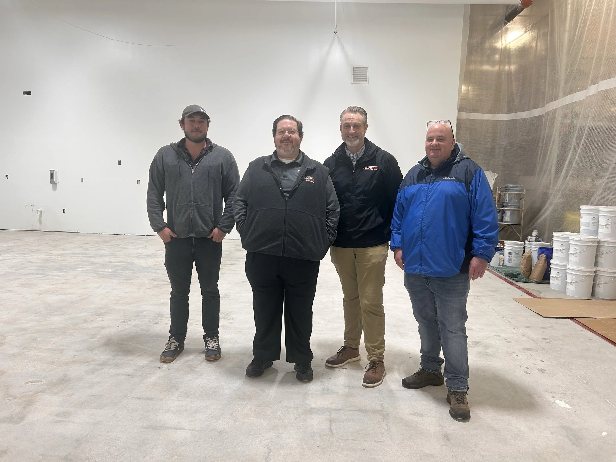 Four men standing in a large, unfinished indoor space, with stacks of white buckets on the right side.