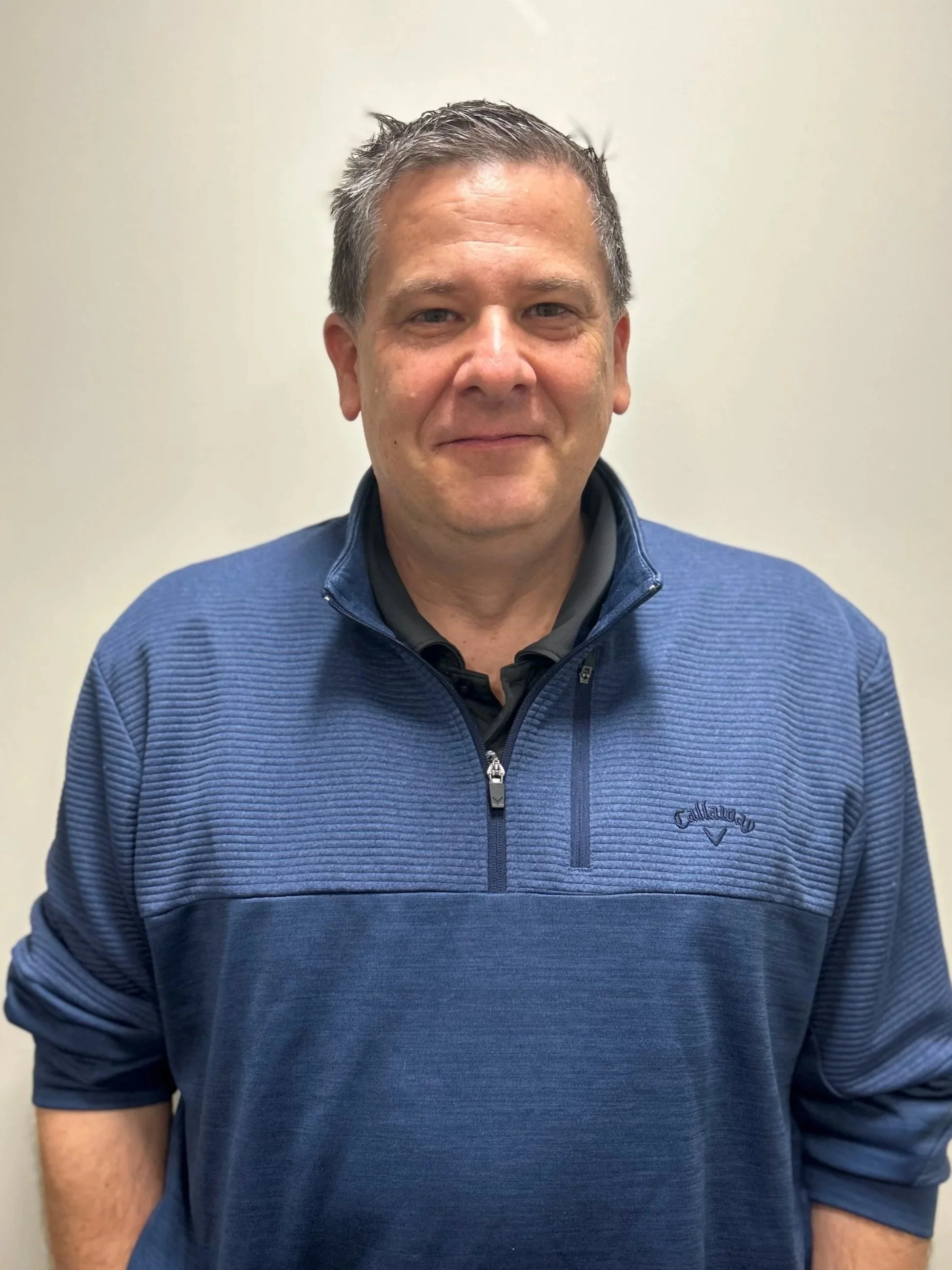 A middle-aged man with short gray hair and a slight smile, wearing a blue zip-up sweater with a Callaway logo, standing in front of a plain, off-white wall.