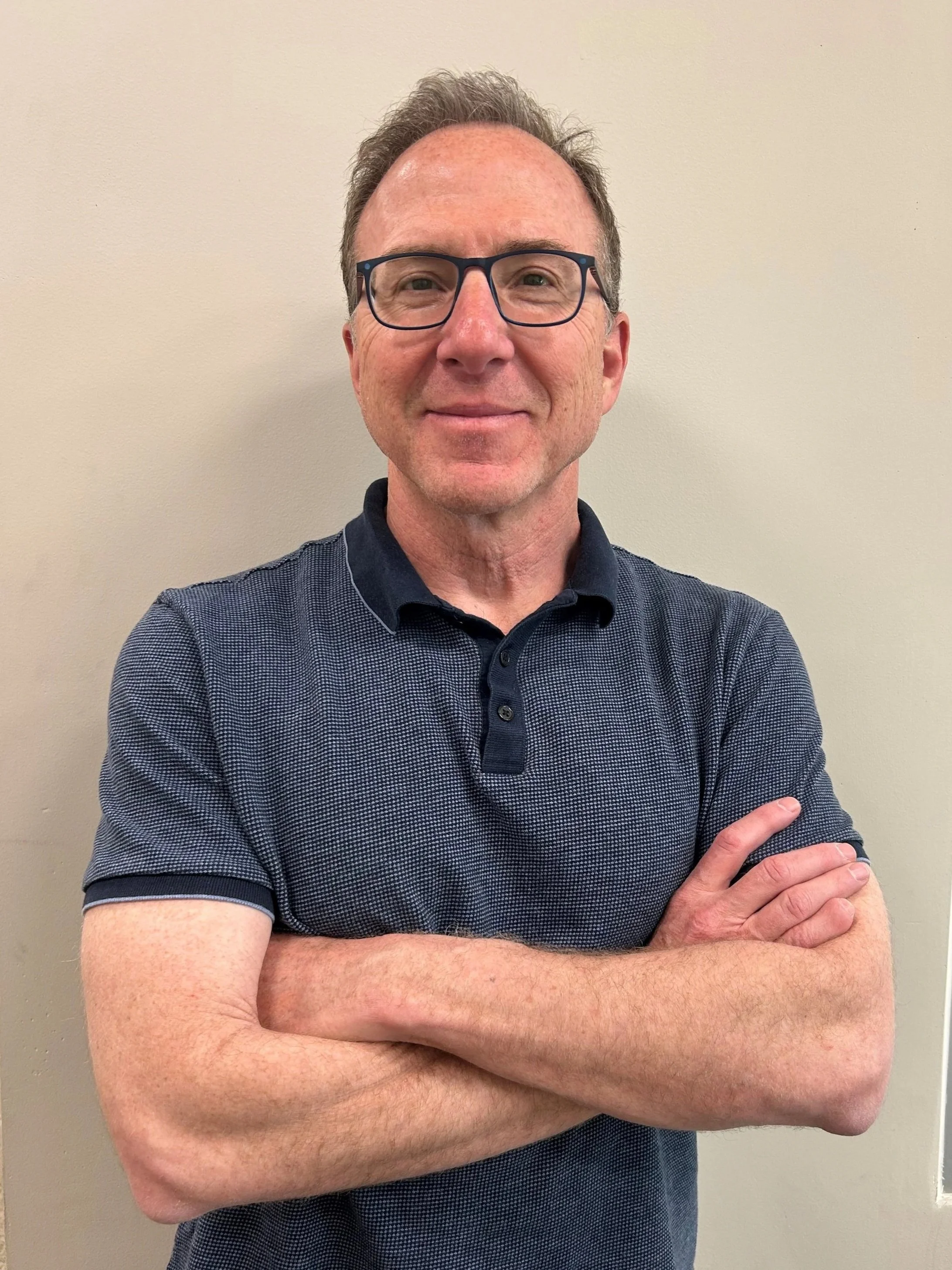 A middle-aged man with glasses, short curly hair, and a slight smile, standing against a plain beige wall with his arms crossed.