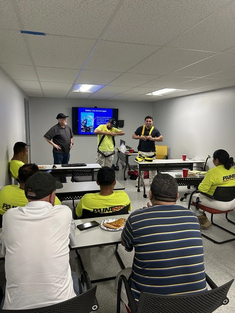 A group of people attending a safety training session in a conference room, with three instructors standing at the front and a presentation slide titled 'Safety Line Anchors' displayed on a monitor.