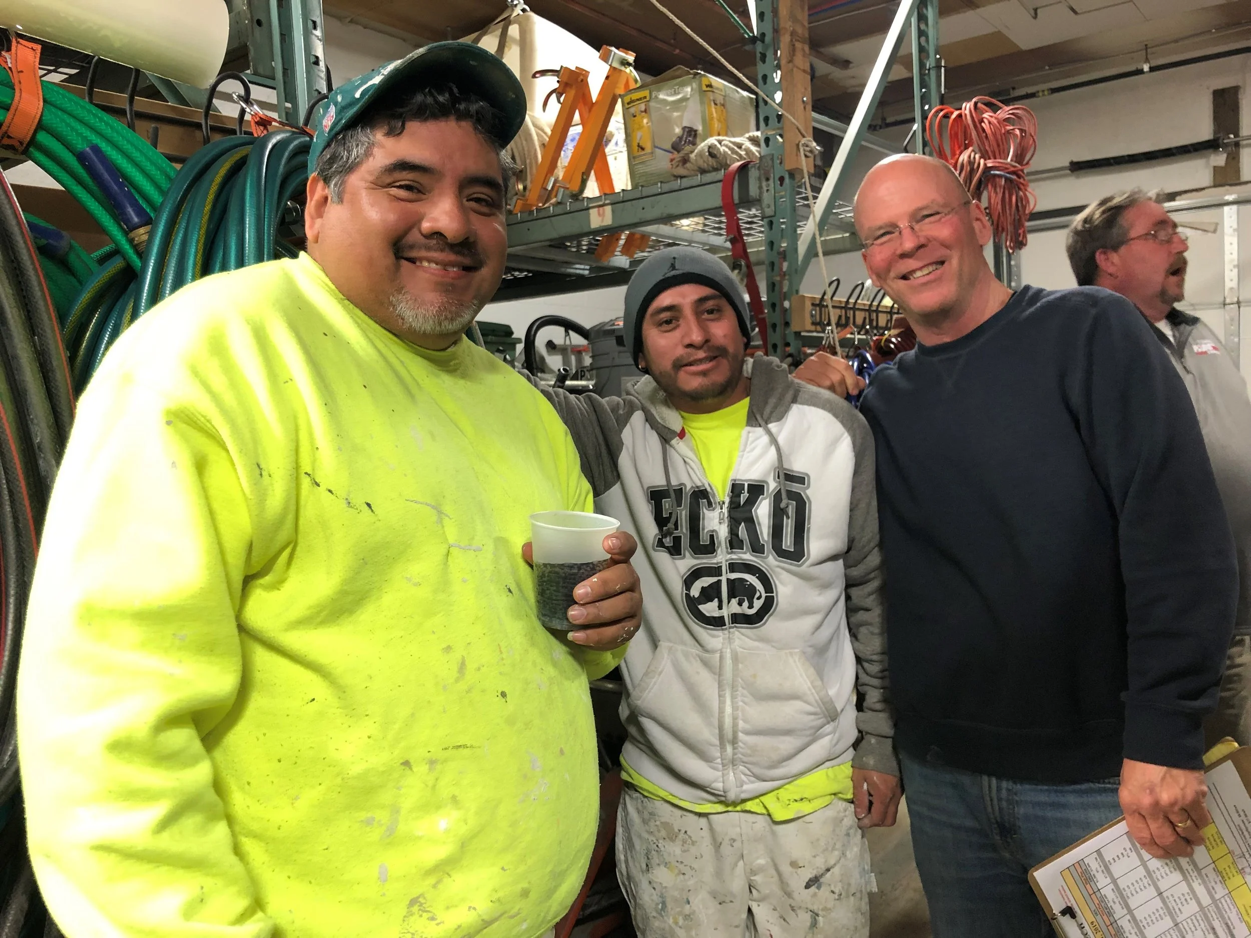 Group of four men standing in a workspace or warehouse, smiling. One man is wearing a bright yellow safety shirt and holding a cup, another is wearing a hoodie and a beanie, the third man is wearing glasses and a dark shirt, and the fourth man is partially visible, holding a clipboard.