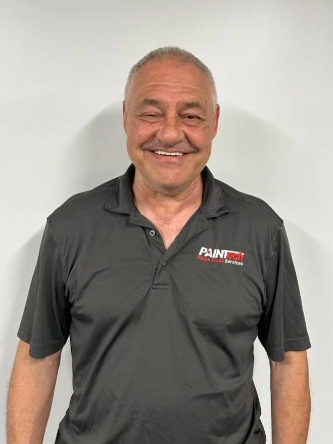 A smiling middle-aged man with short gray hair, wearing a dark gray polo shirt with a logo that reads 'PAINtech Florida Trade Services,' standing against a plain white wall.