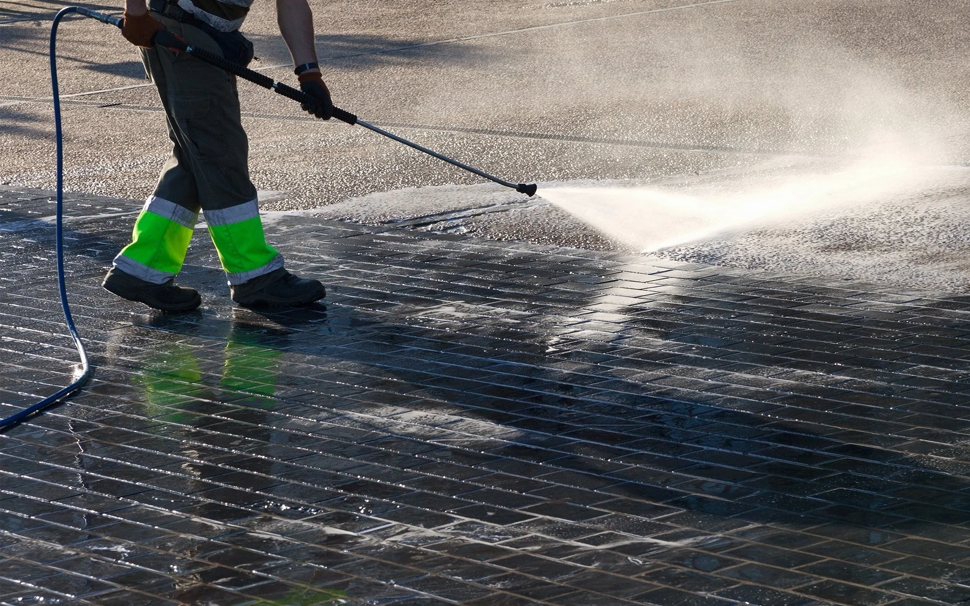 Worker pressure washing a large metal grate outdoor, creating water spray and steam.