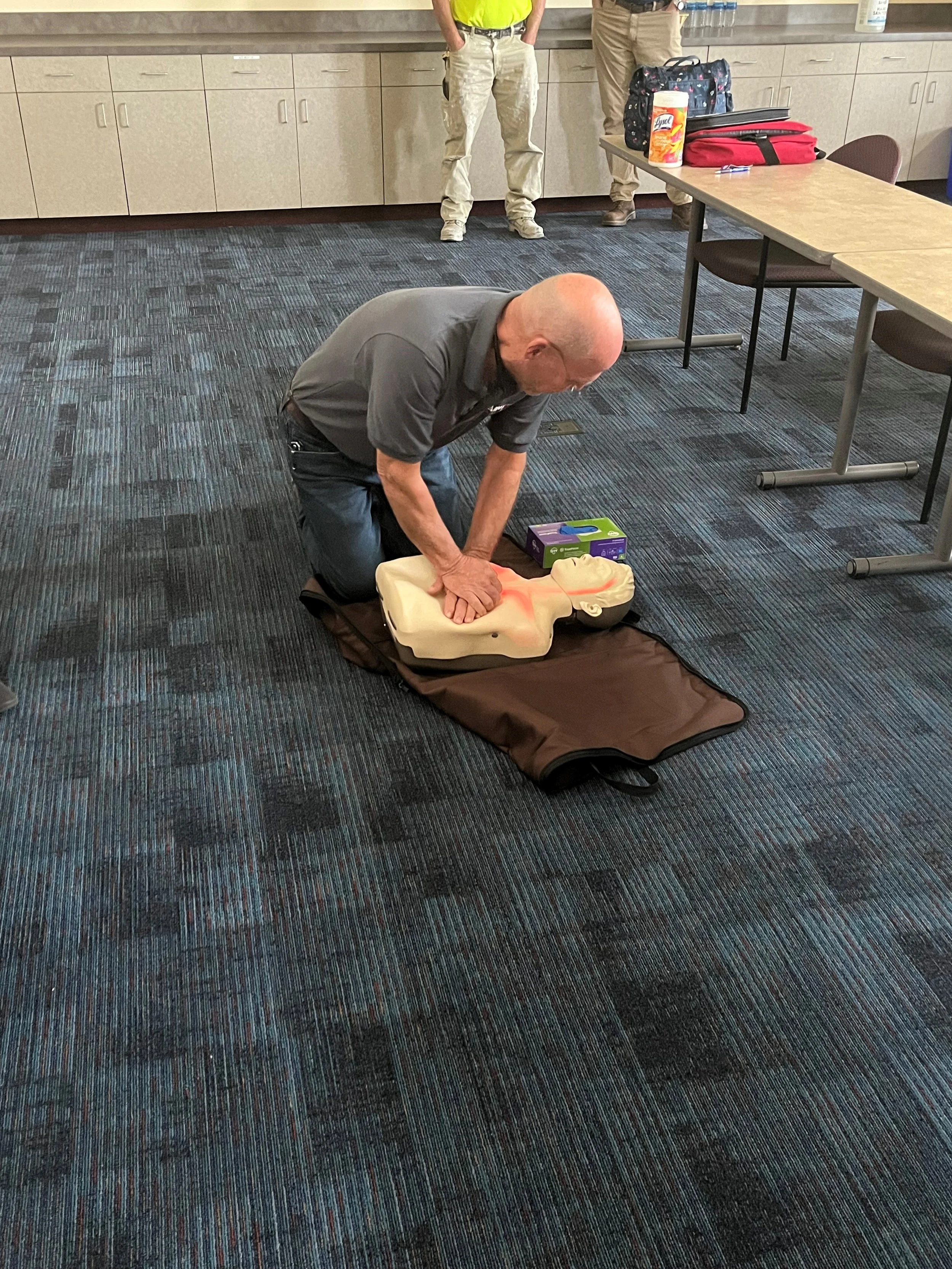 A man performing CPR on a training mannequin on a carpeted floor, with two other people standing behind him in a room with cabinets and tables, supplies for a training session nearby.