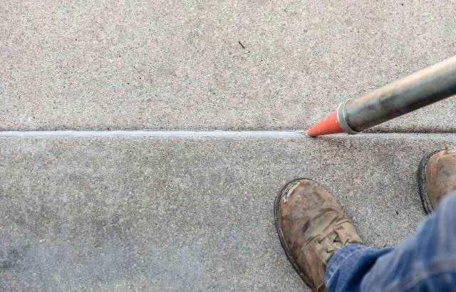 Person wearing brown work boots standing on concrete sidewalk next to a sidewalk chalk line drawn with orange chalk, with a gray metal tool nearby.