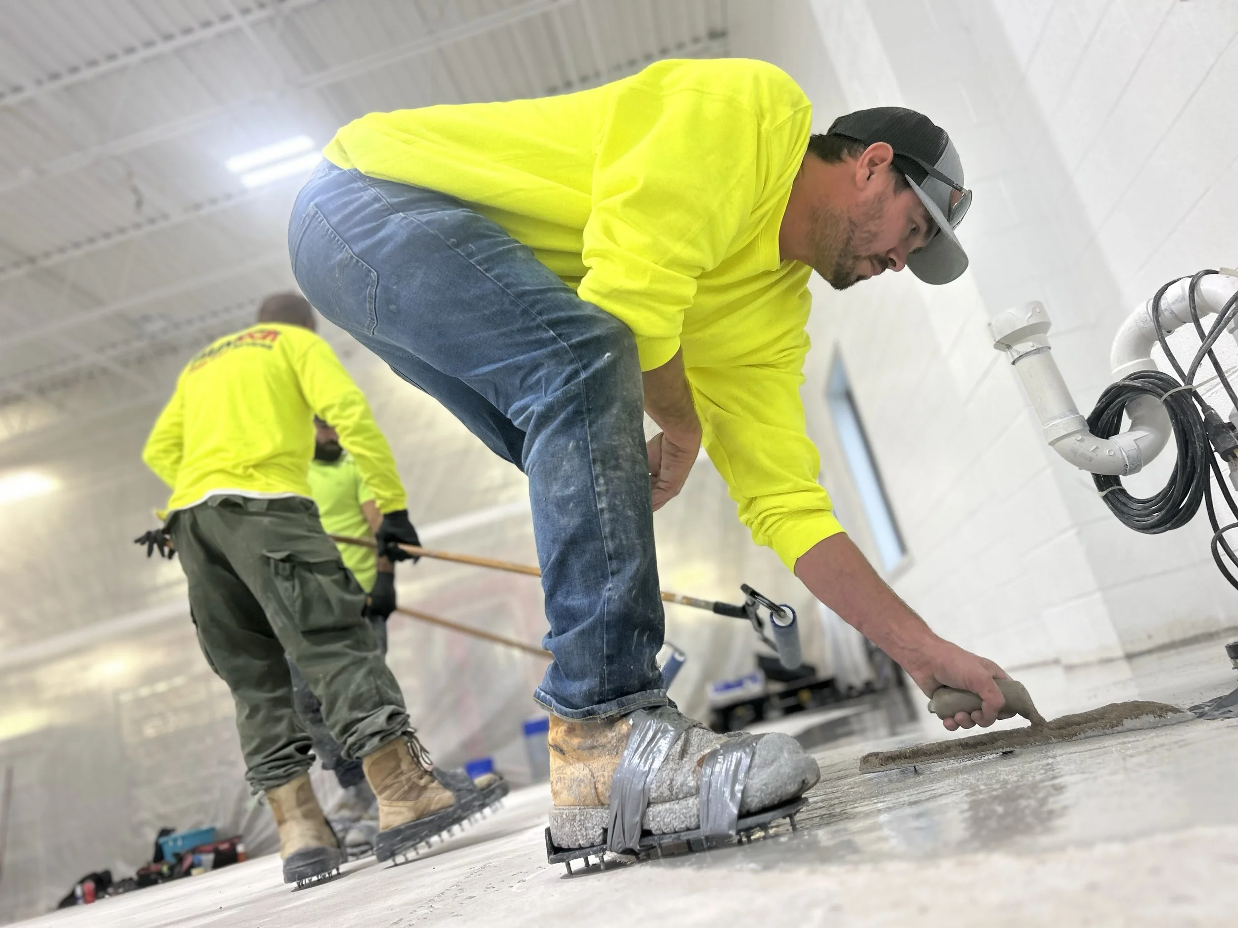 Construction workers in high-visibility yellow shirts working on a floor, with one worker kneeling and using a trowel while another stands in the background, all inside a building with a ceiling and pipes.