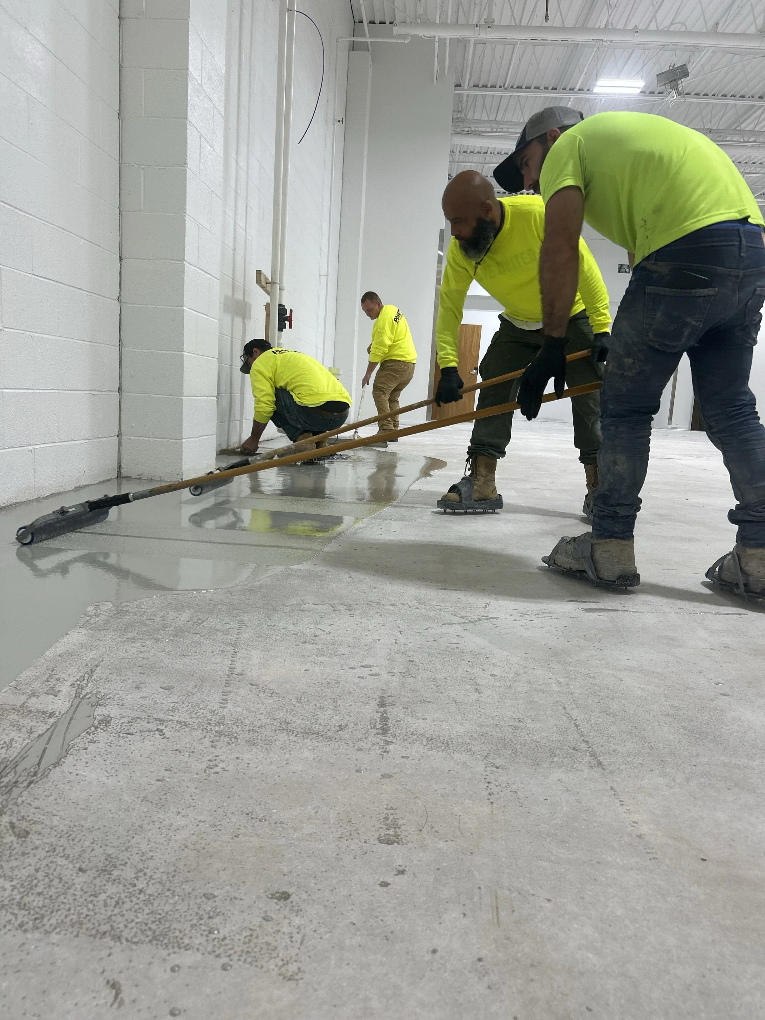 Four workers in bright yellow shirts applying a layer of white epoxy coating on a concrete floor in an industrial or commercial space.