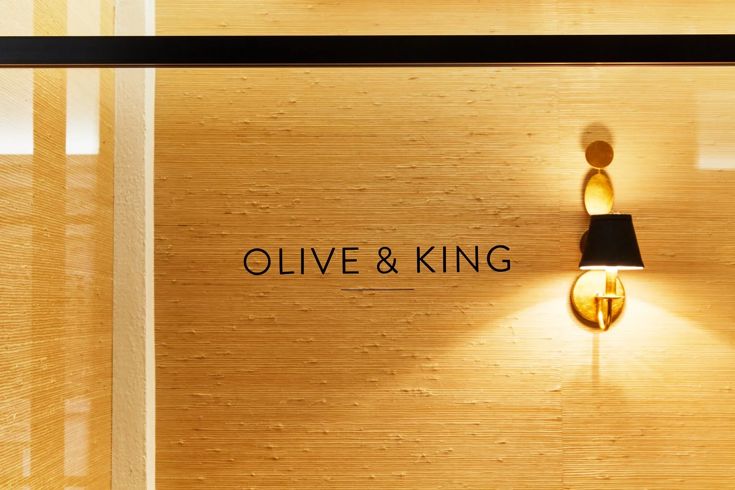Interior wall with textured beige surface, a modern gold sconce with a black lampshade, and the words "OLIVE & KING" in black text.