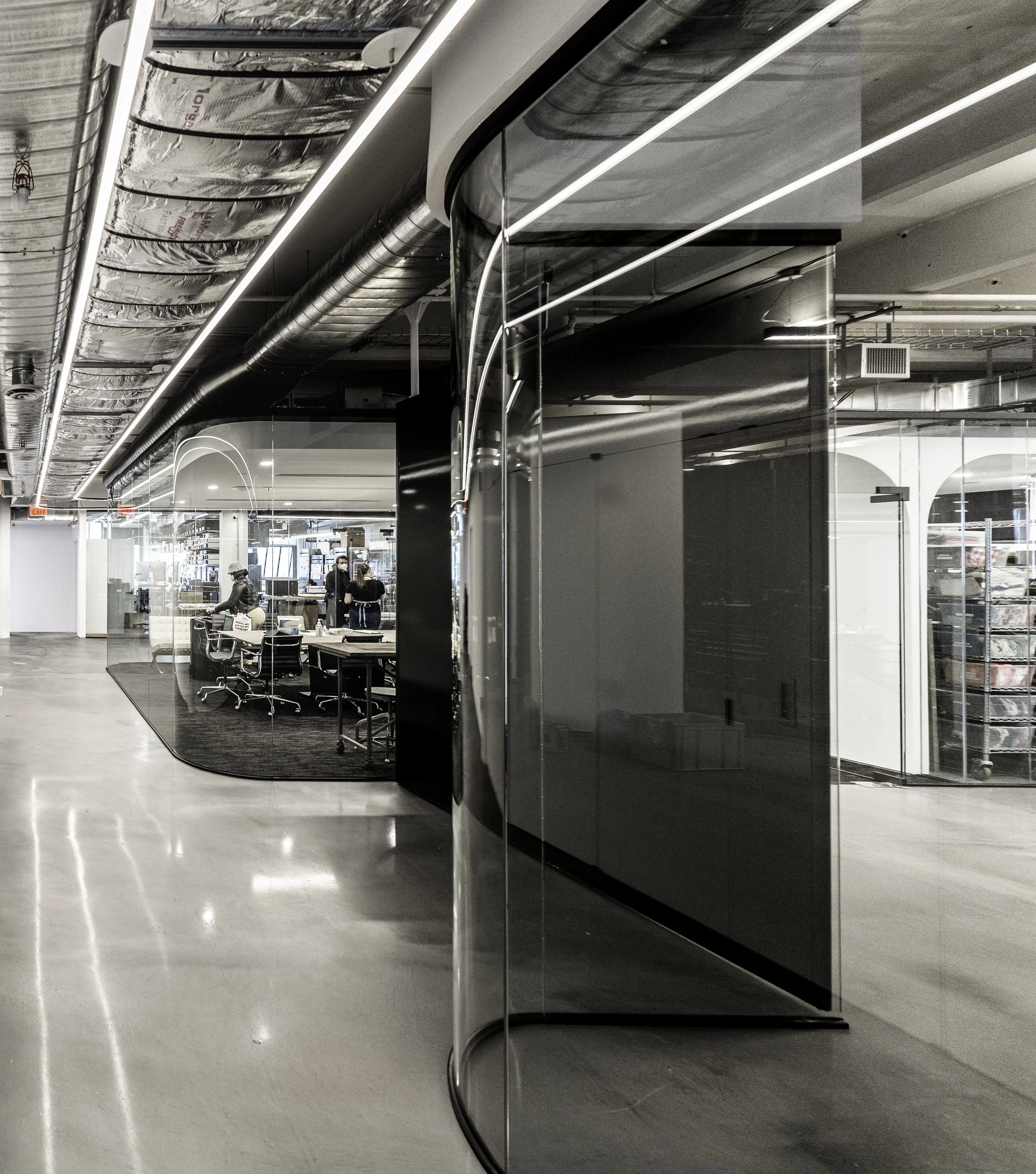 Modern office interior with glass walls, exposed ceiling ductwork, and people working at desks.