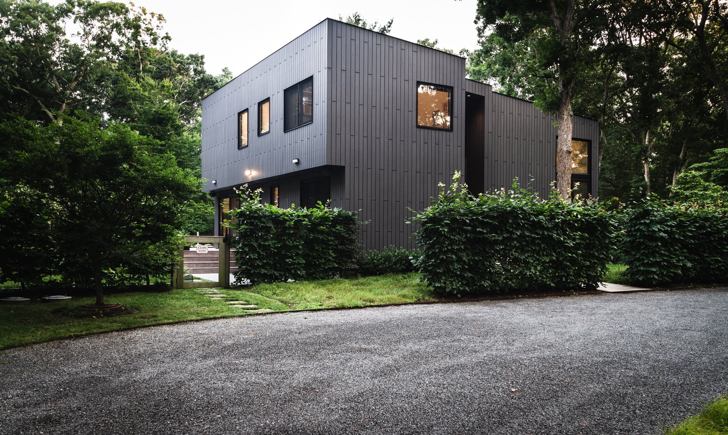 Modern two-story house with dark cladding, surrounded by trees and shrubs, a gravel driveway in front.